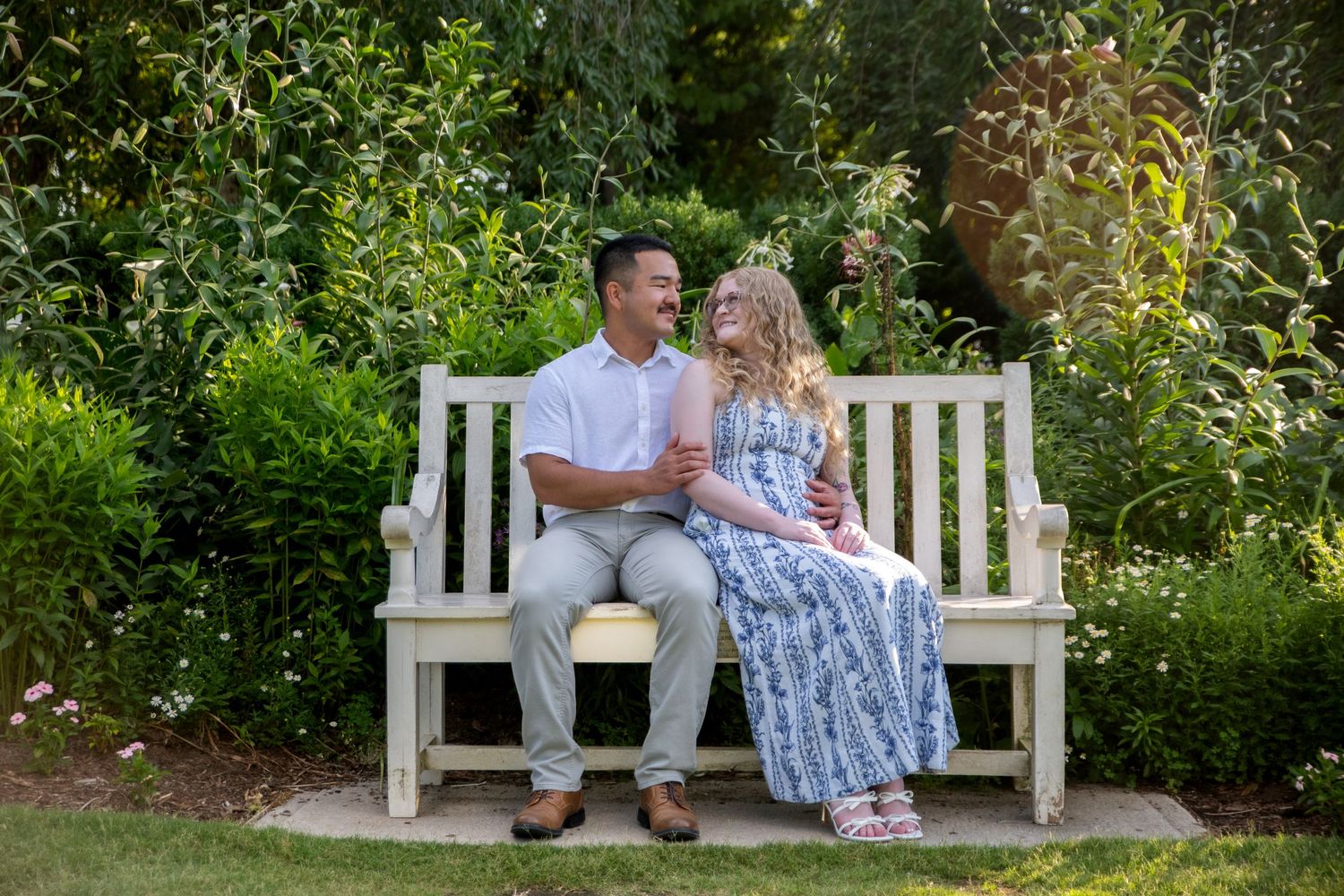 Happy couple sitting together on a white wooden garden bench surrounded by lush green trees.