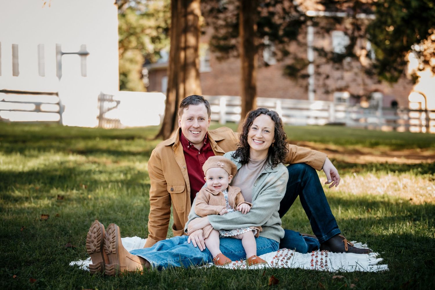 Two women and a toddler smile together on a blanket in a sunny green park during a family photo session.