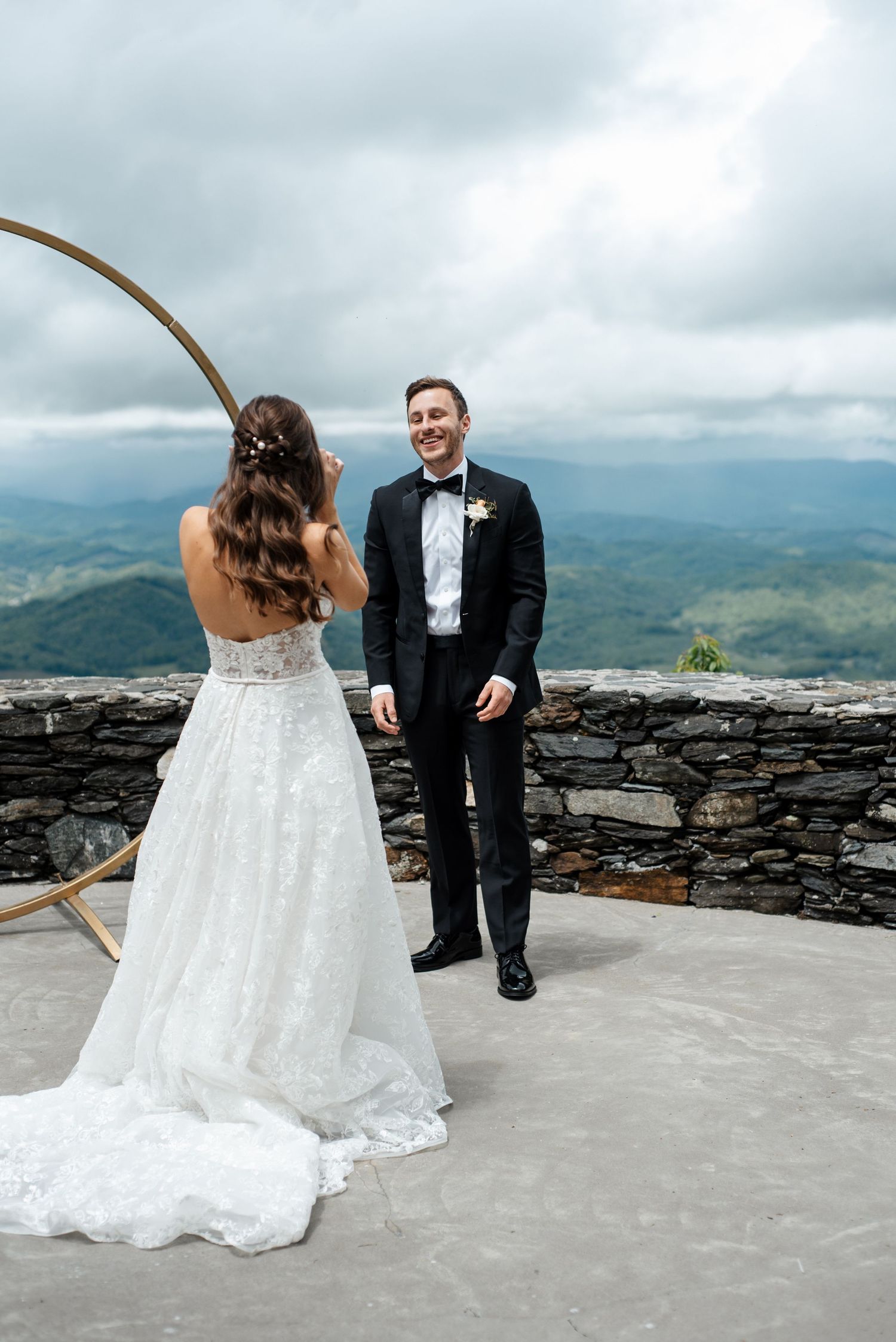 Bride and groom share first look on mountain overlook terrace with circular gold arch and cloudy sky backdrop.