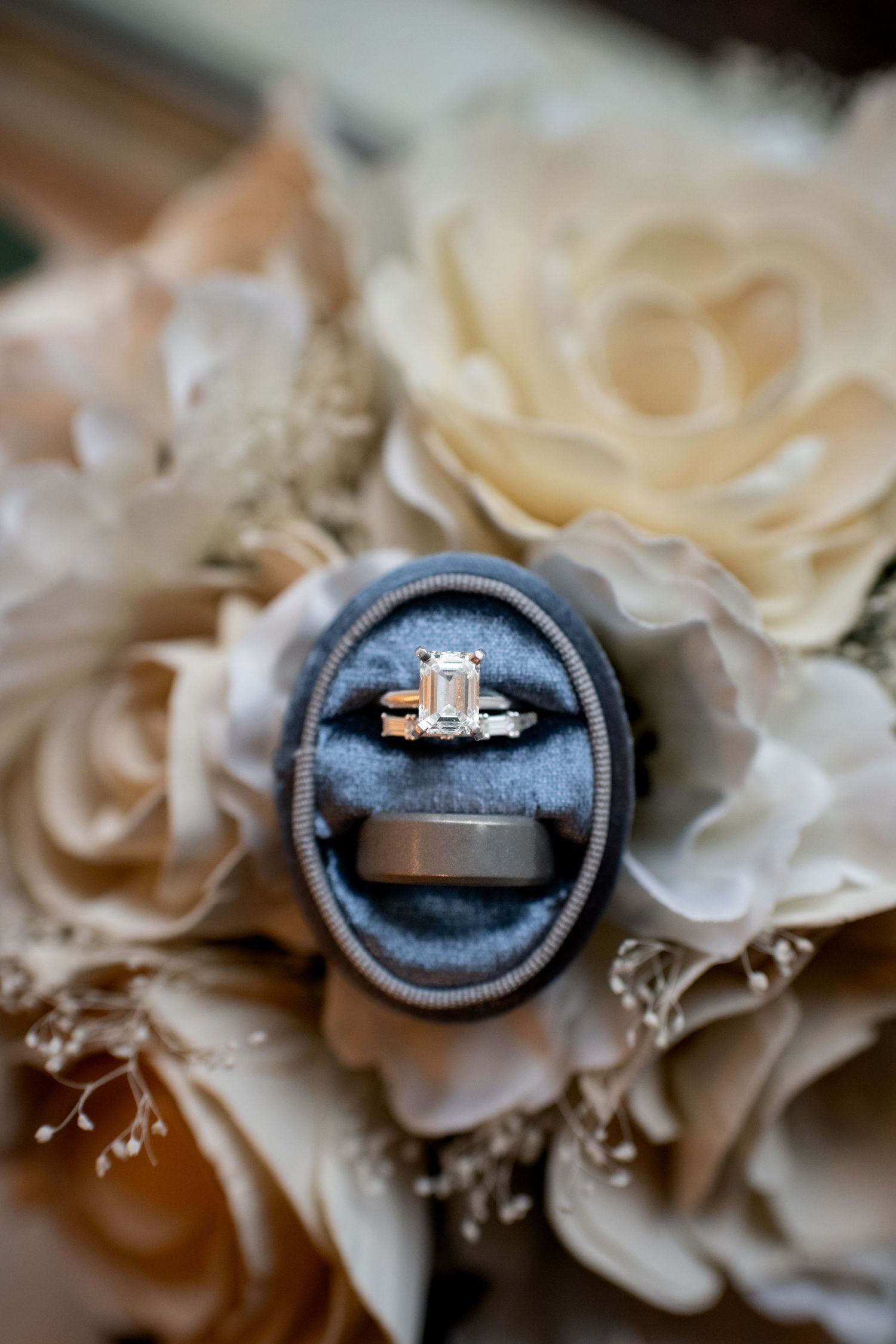 An engagement ring nestled in a navy blue ring box surrounded by cream-colored flowers.