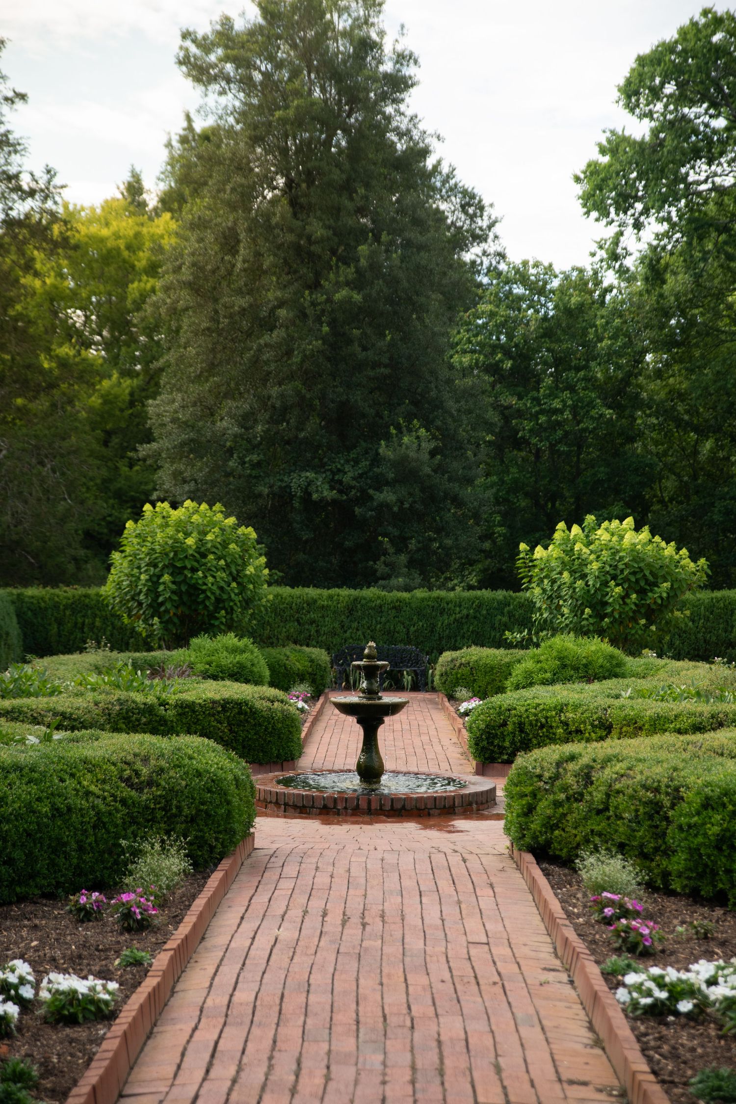 Formal garden with brick pathway leading to tiered fountain, surrounded by manicured hedges and lush green trees.