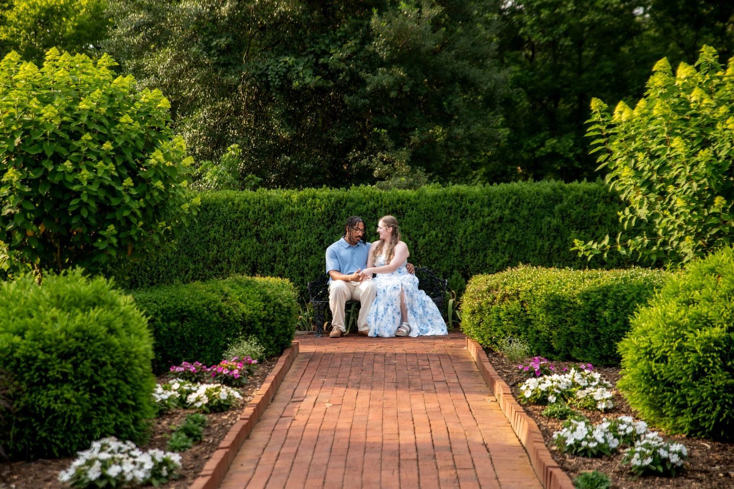 Couple sitting together on a brick pathway surrounded by lush green hedges and colorful flowers in a formal garden.