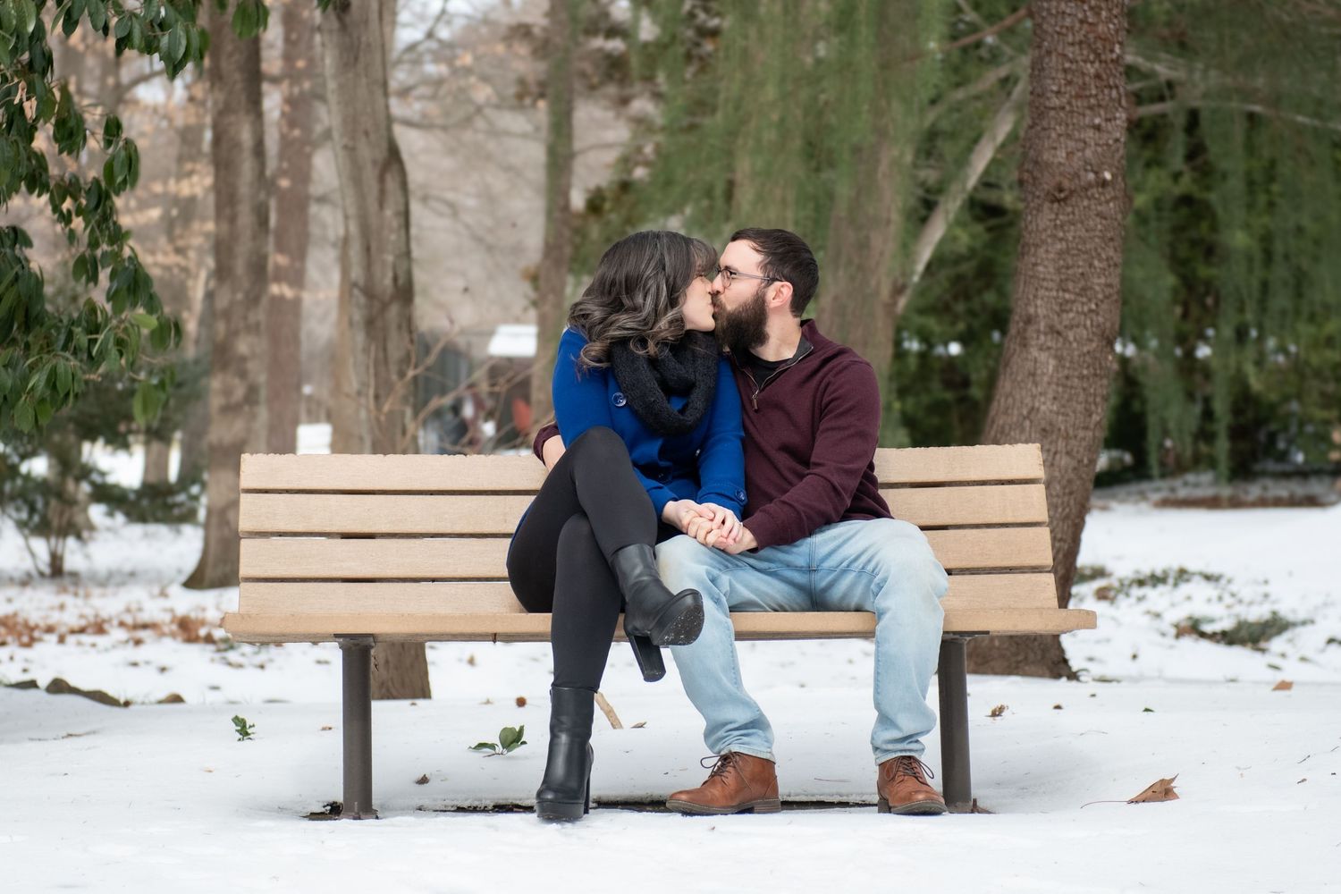A couple sits together on a wooden bench in a snowy park setting during winter.