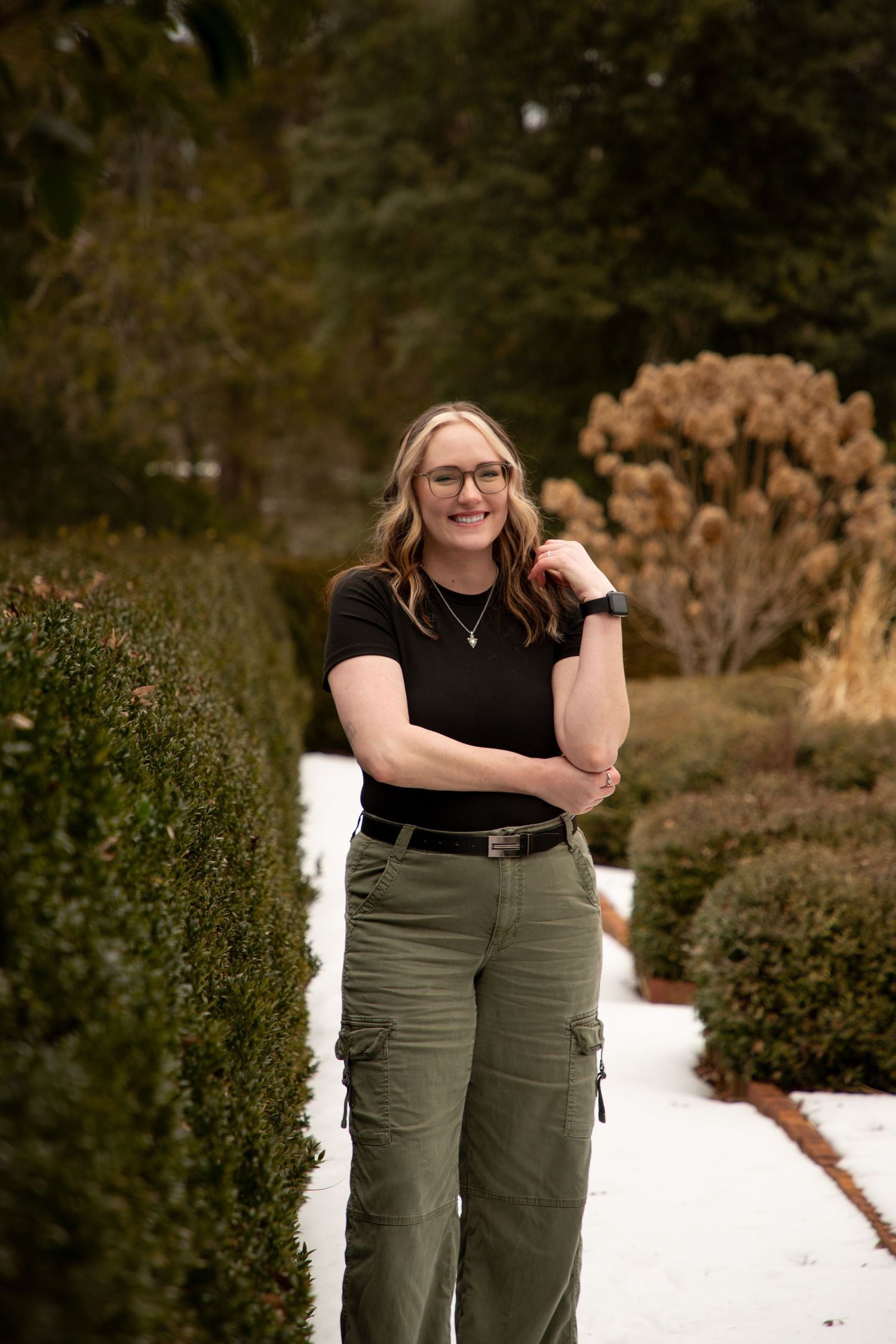 A person in a black top and olive cargo pants poses outdoors in a garden setting with natural fall foliage.