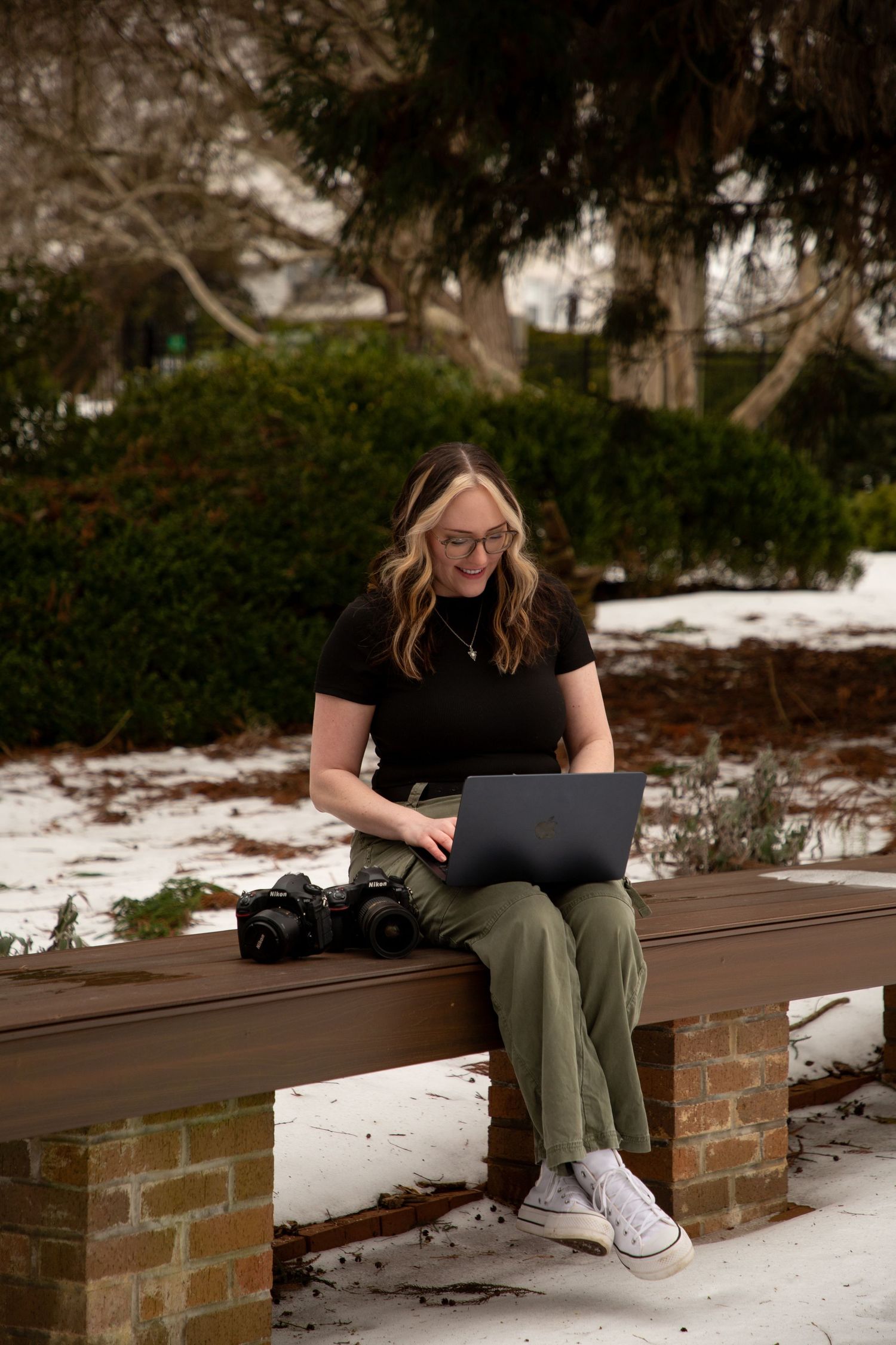 A person in olive pants and white sneakers sits on a brick wall with a laptop while snow covers the ground.