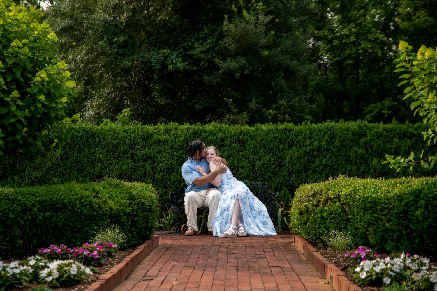 Couple sharing a romantic embrace on a brick garden path surrounded by lush green hedges and colorful flowers.