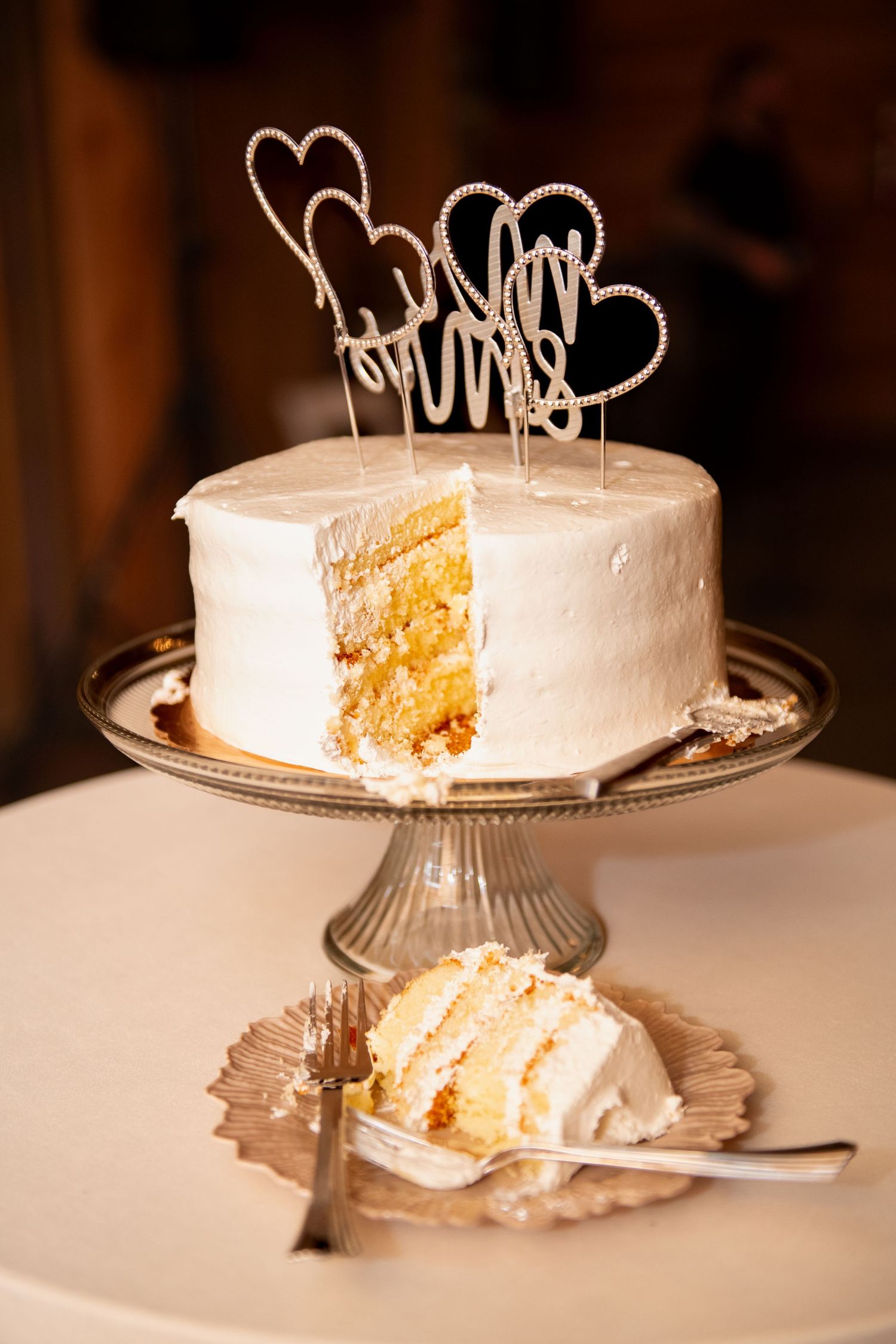 White wedding cake with rhinestone heart toppers on glass stand, with a slice cut and plated beside it.