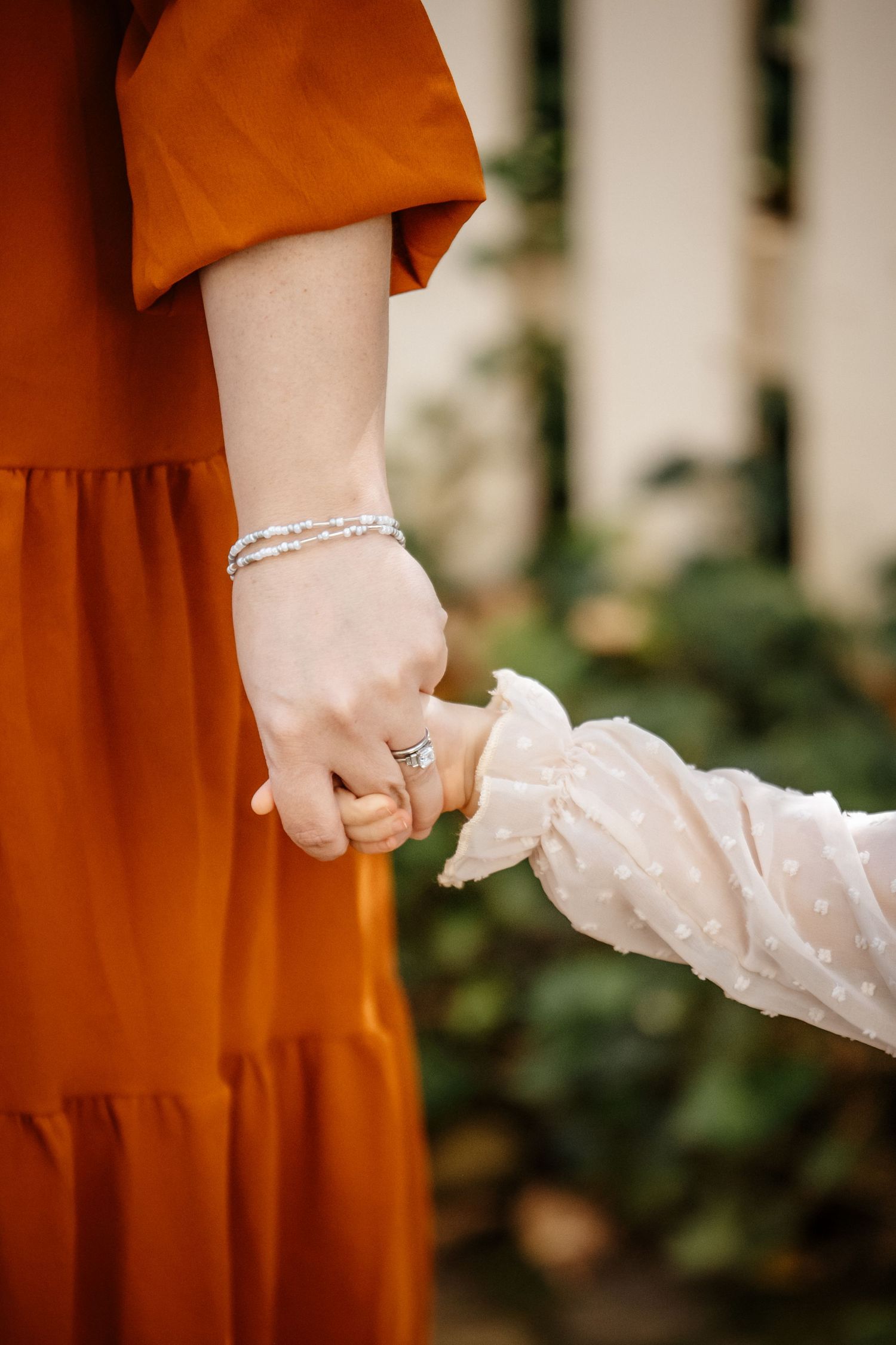 A person wearing a rust-colored dress and white lace sleeves stands with their hands clasped together in an outdoor setting.