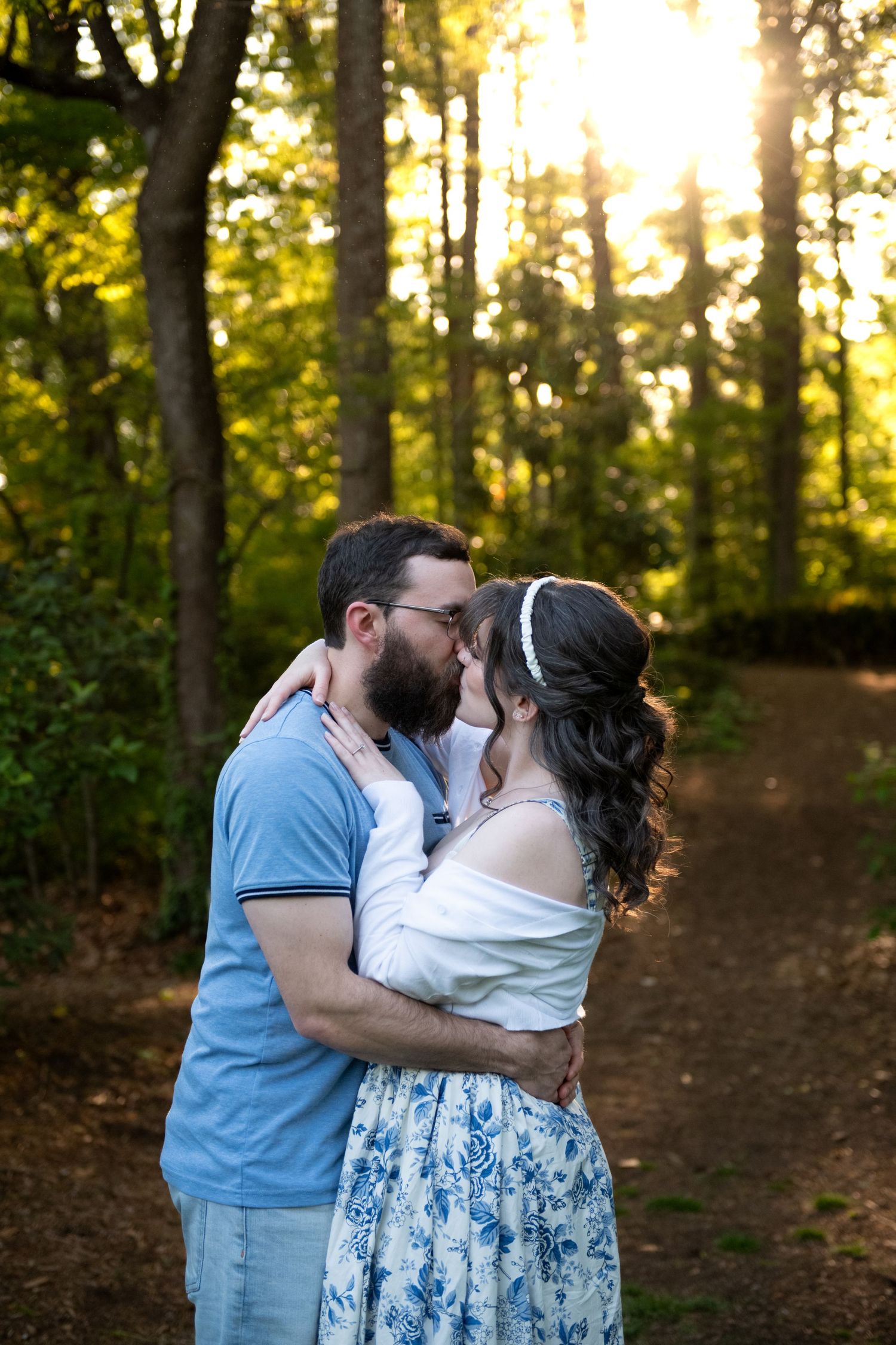 Couple sharing a romantic kiss on a sunlit forest path, woman in floral skirt embracing bearded man in blue polo.