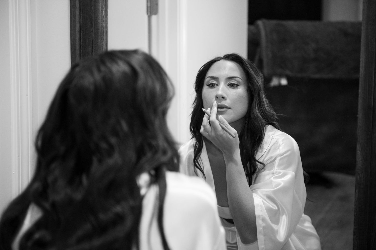 Black and white photo of a woman in a silk robe applying lipstick while looking in a mirror.