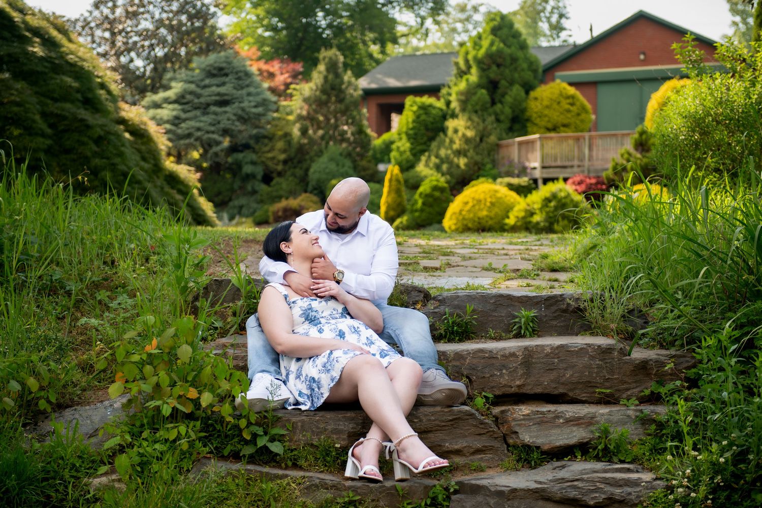 Couple sitting on stone steps in a lush garden, sharing a tender moment with a rustic building in the background.