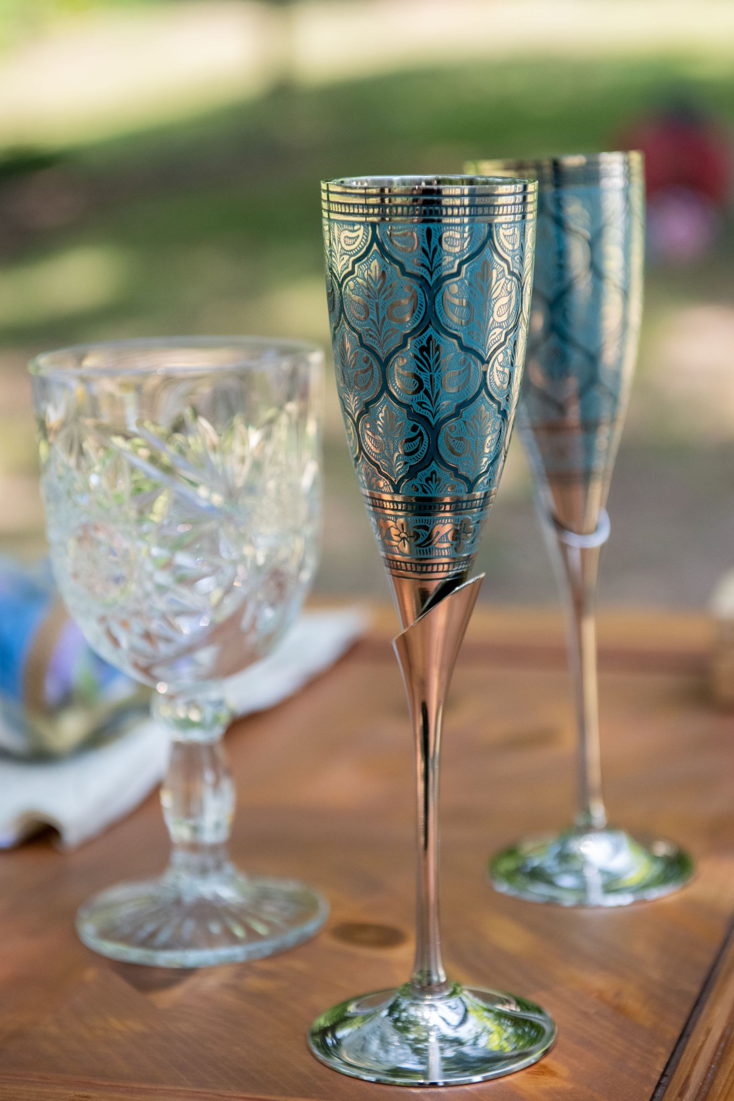 Elegant decorative champagne flutes with teal and gold patterns beside a crystal wine glass on a wooden table outdoors.