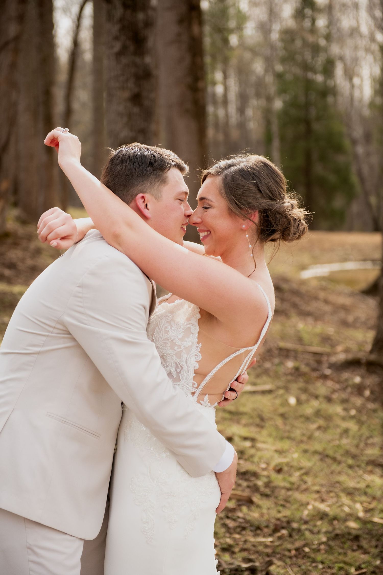 A joyful bride and groom embrace outdoors, laughing together, she in a backless lace gown, he in a light gray suit.