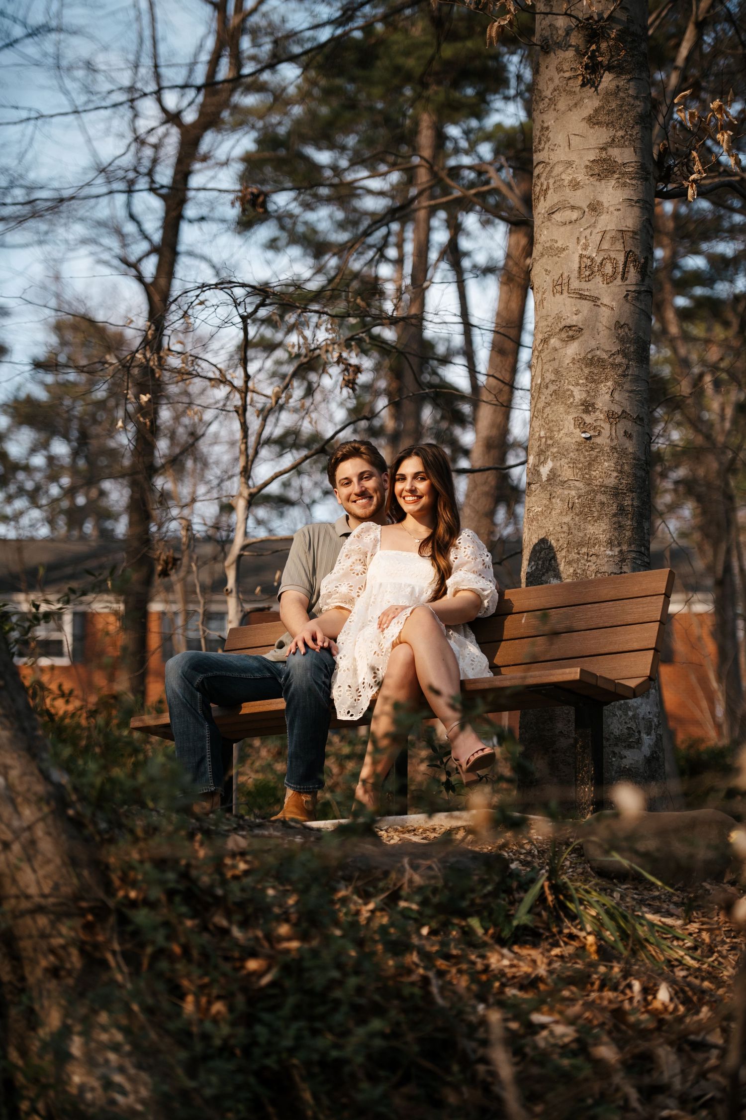 A couple relaxes together on a park bench surrounded by tall trees in autumn.