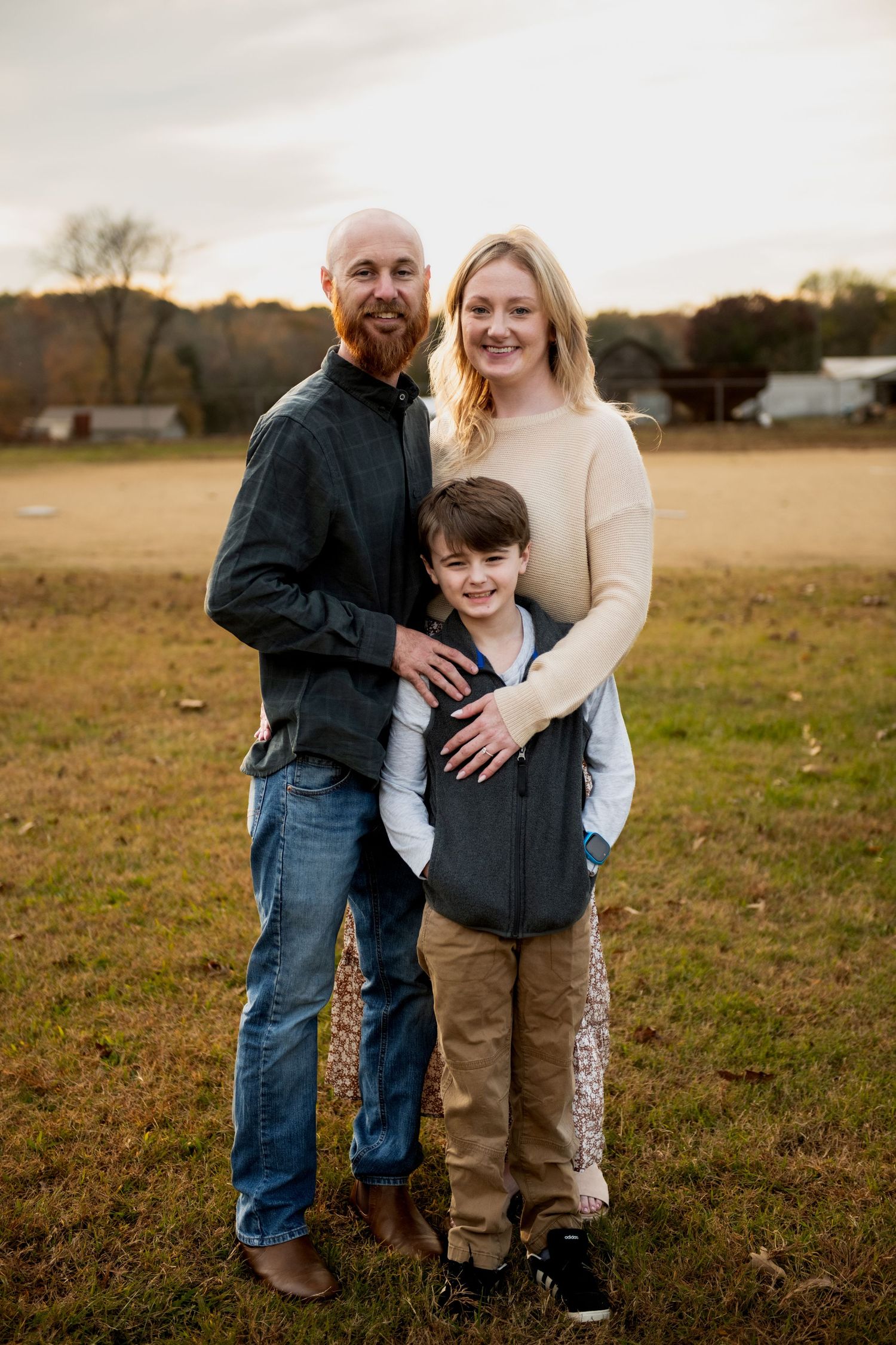 A family of three poses outdoors in a field during autumn, with a bald bearded man, blonde woman, and young boy smiling.