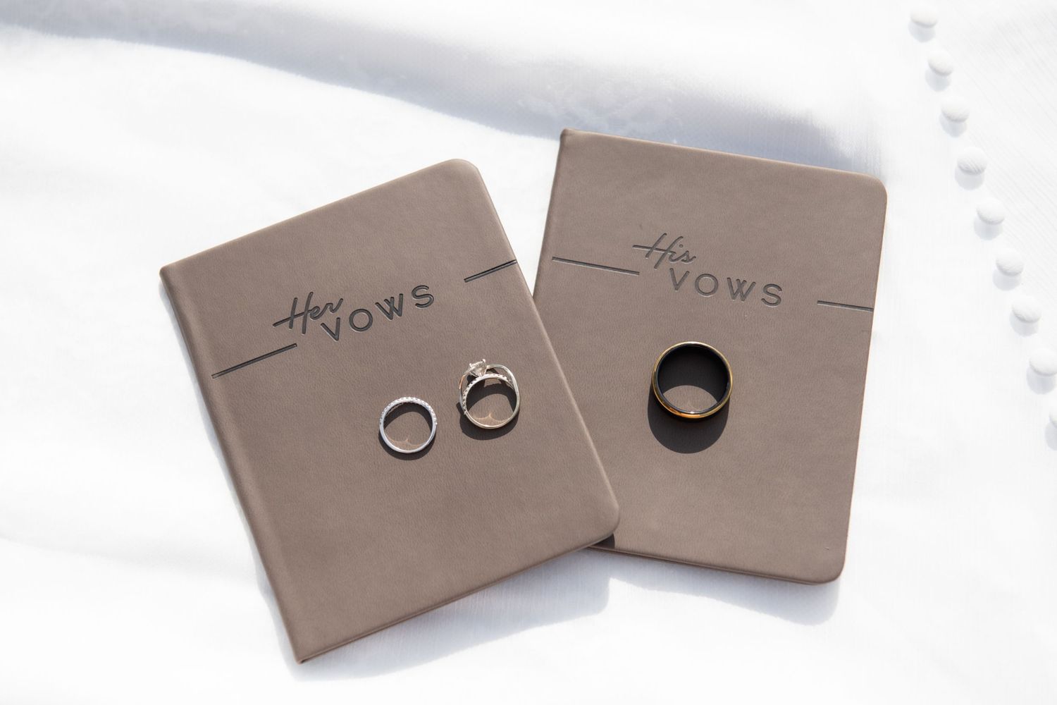 Two wedding vow books with his and hers rings resting on top, displayed on a white fabric background.