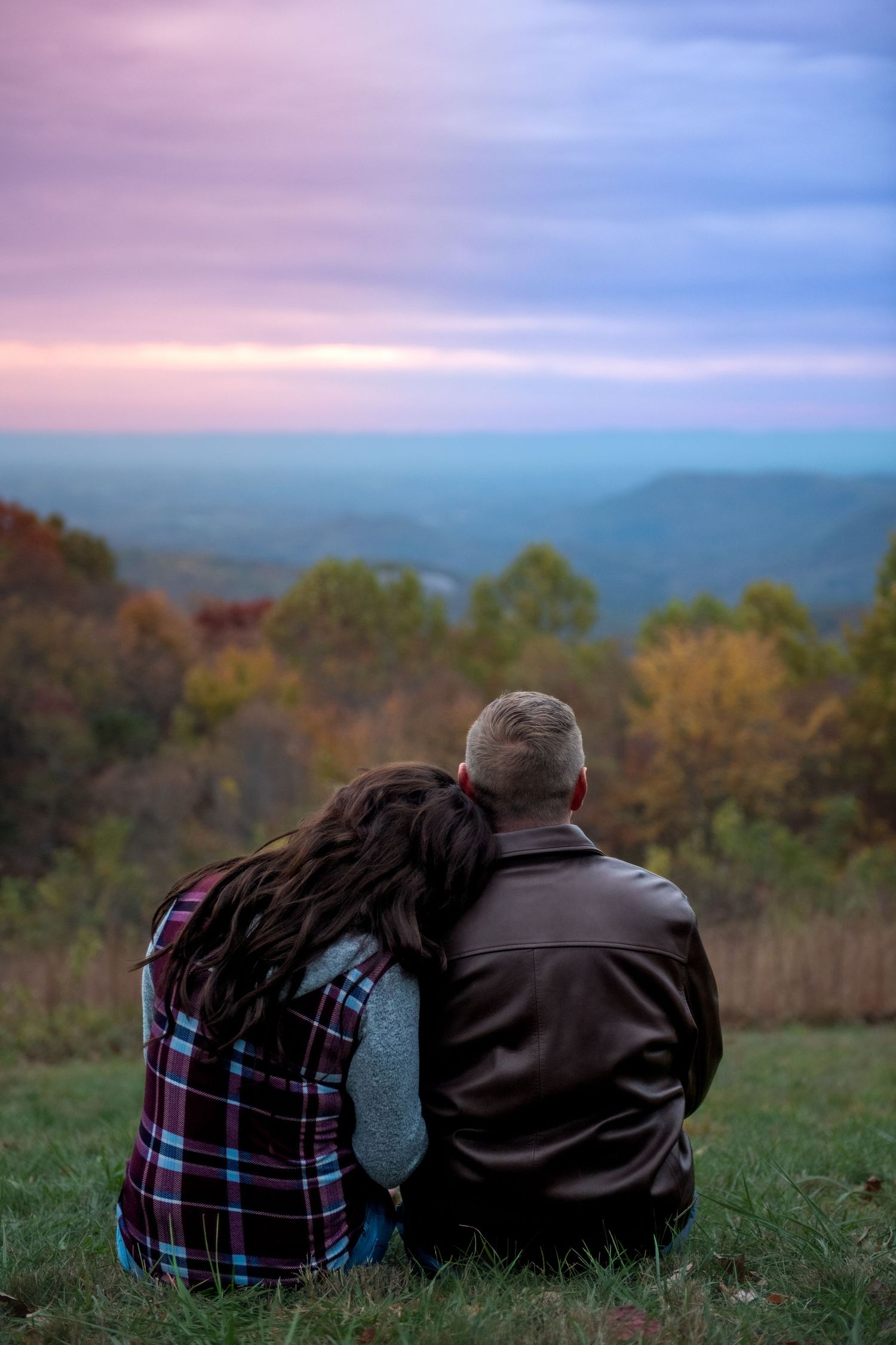 Two people in warm clothing sit together on a grassy hillside watching a colorful sunset over mountain scenery.