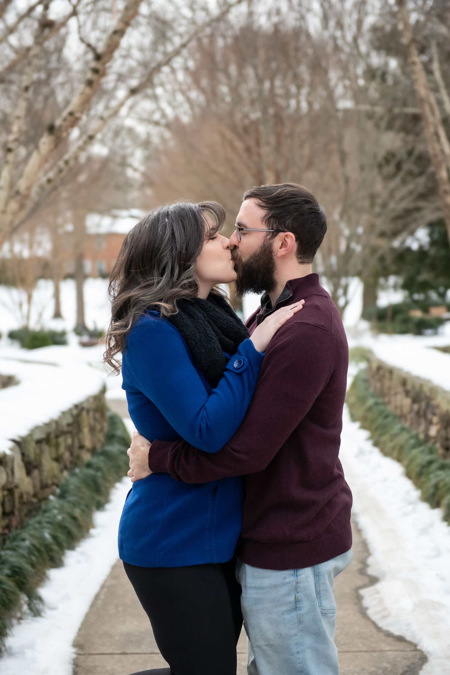 A couple shares romantic kisses while standing on a snowy winter path lined with bare trees.