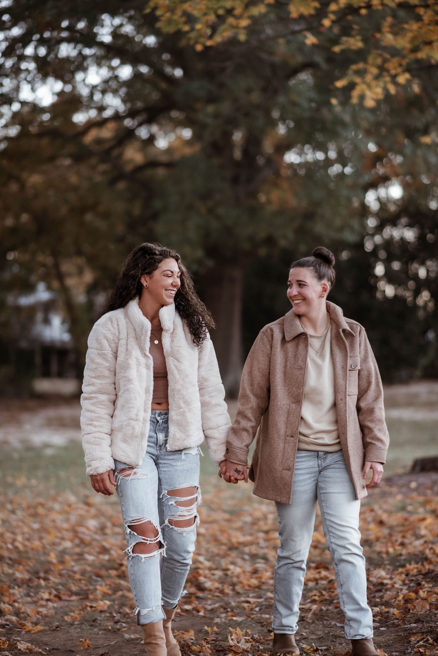 Two people in casual outfits share laughter while walking through autumn leaves in a park setting.