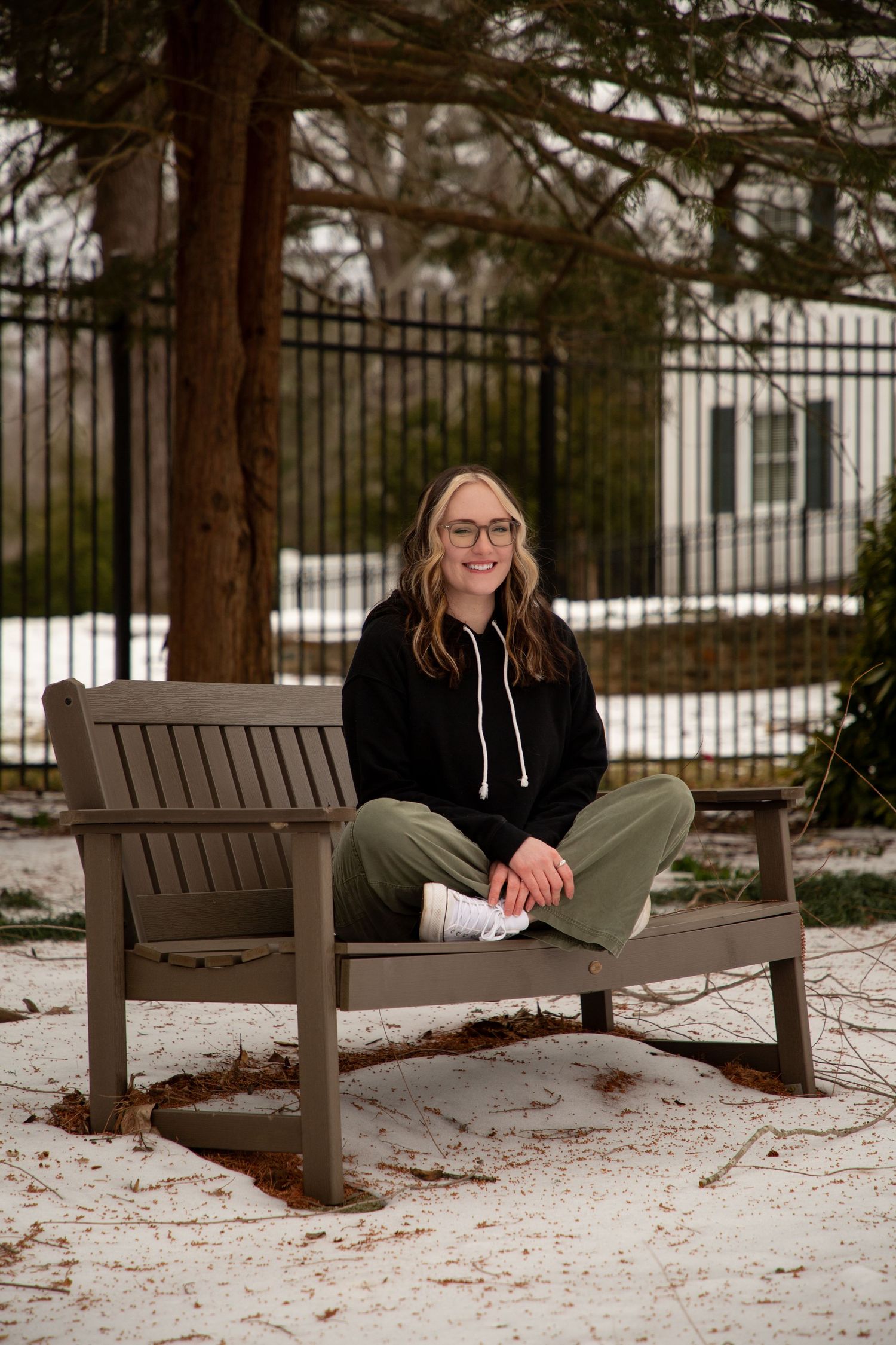 Someone in a black hoodie and glasses sits casually on a brown bench in a snowy outdoor setting with trees and fence.