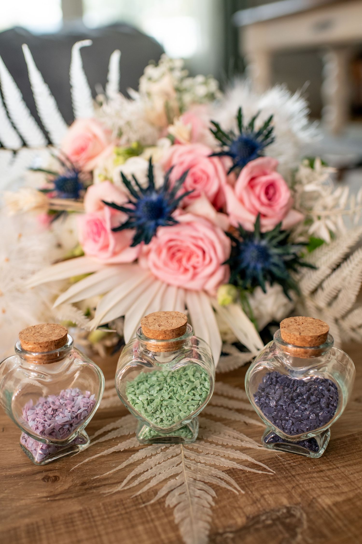Decorative glass bottles filled with colorful sand displayed alongside pink roses and navy blue flowers.