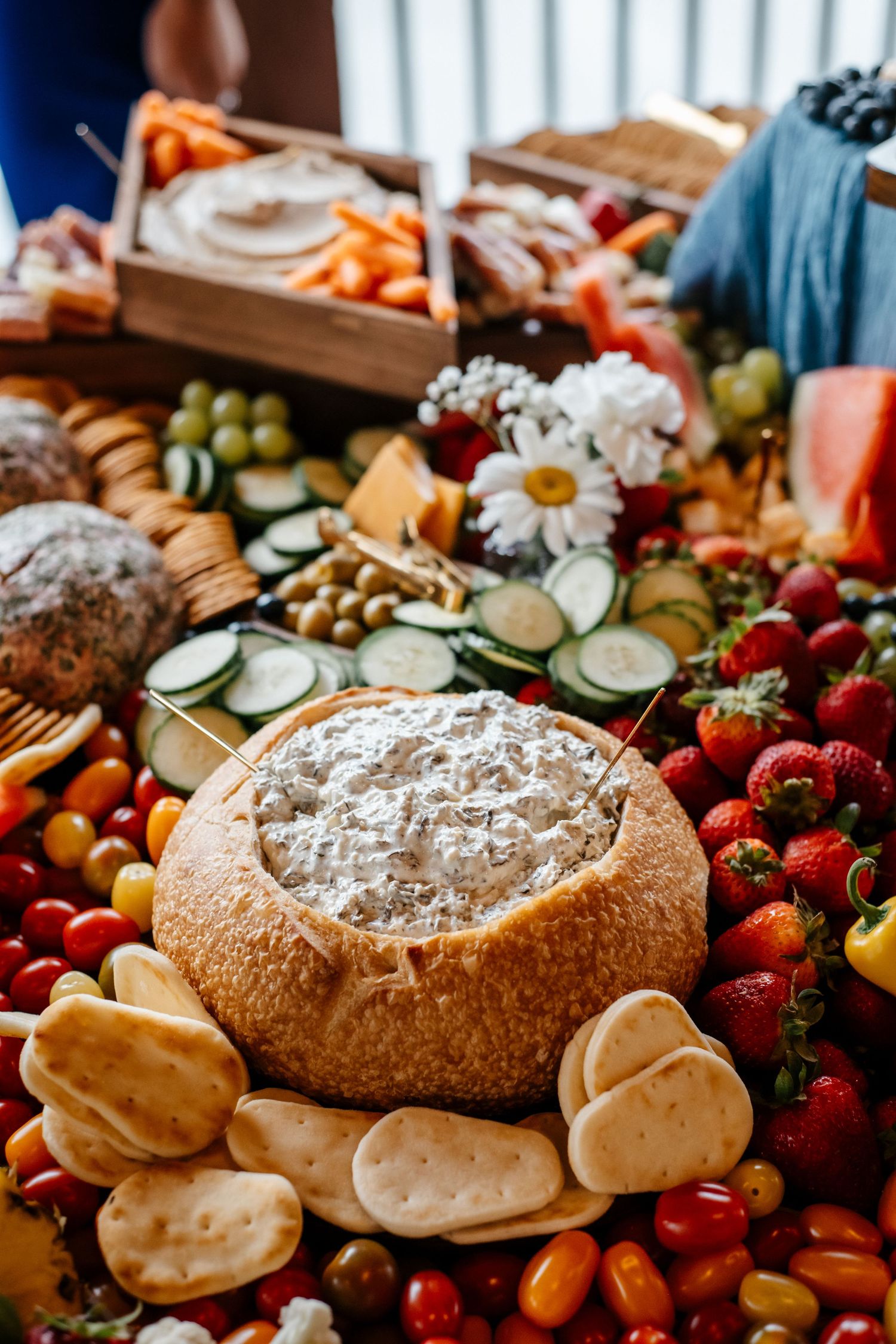 Elegant charcuterie board with bread bowl dip, crackers, fresh strawberries, tomatoes, cucumbers, grapes, and flowers.
