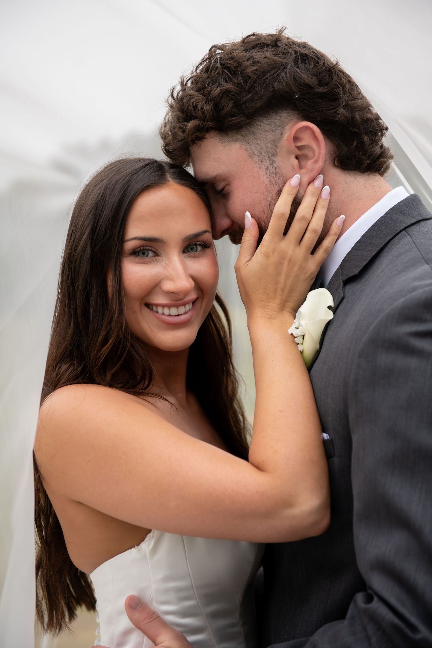 A smiling bride in a white dress tenderly holds a groom's face as they share an intimate moment at their wedding.