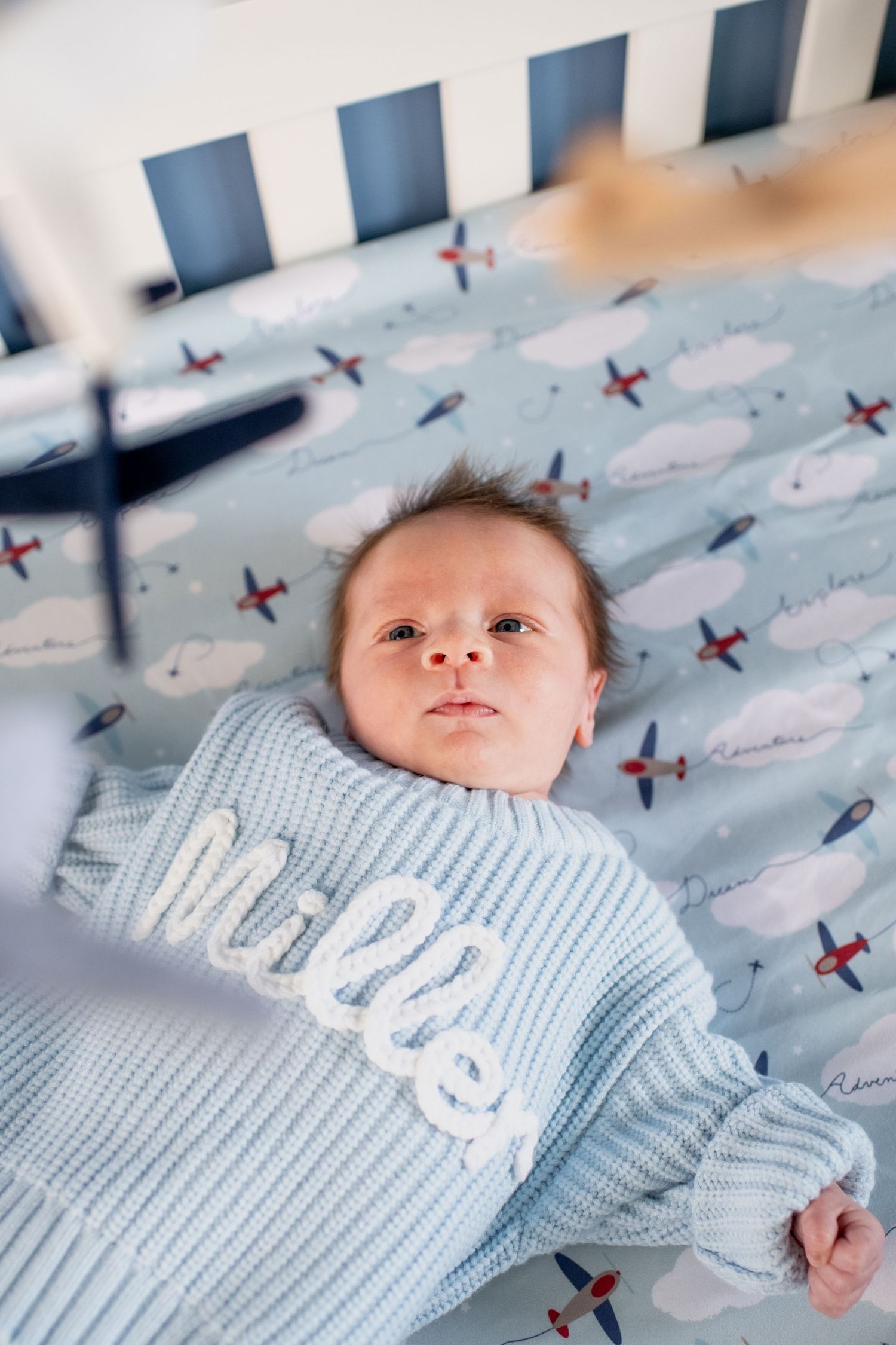 Baby wearing light blue knit sweater lies on airplane-patterned bedding in a crib.