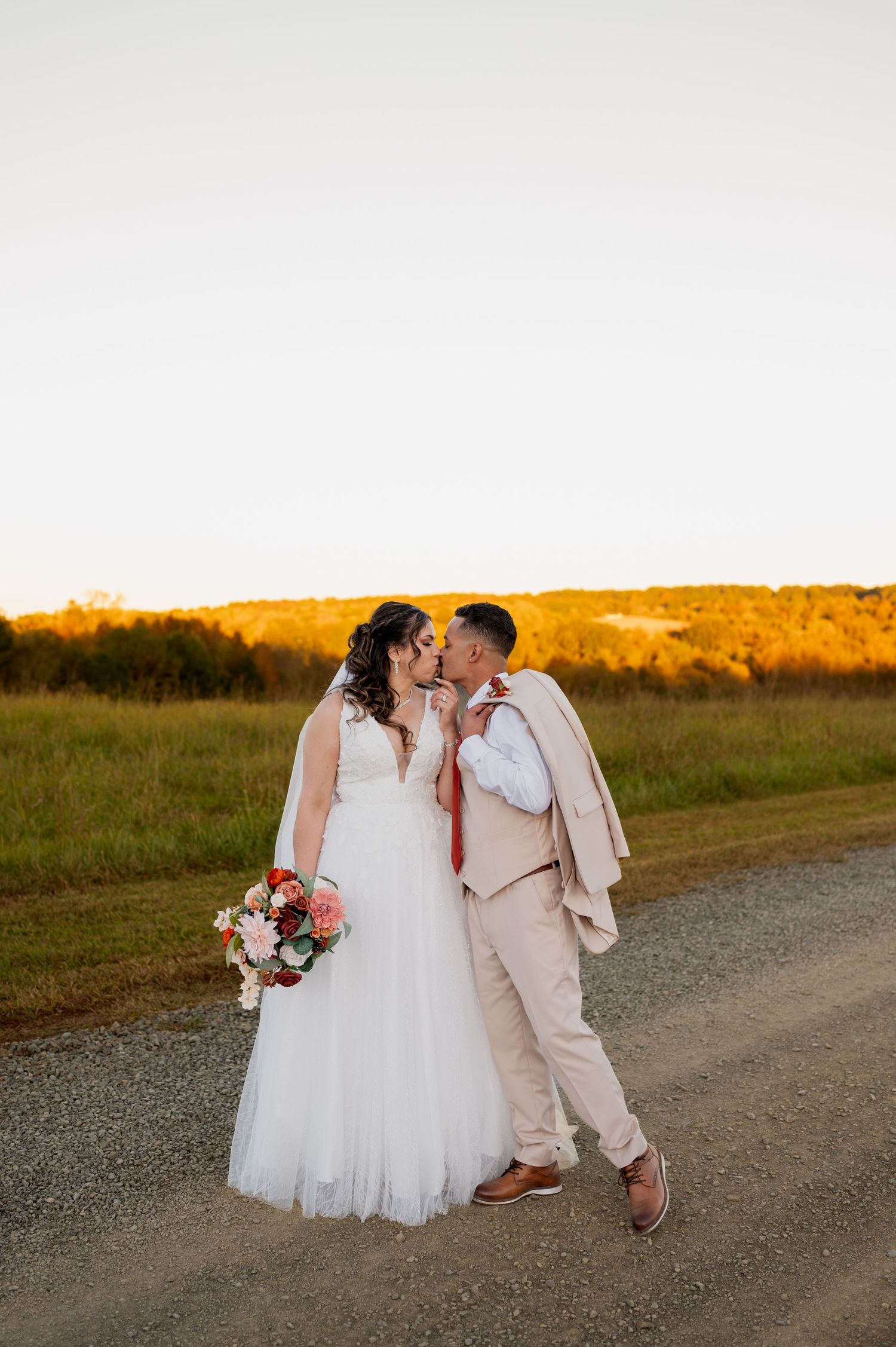 A couple walks together on a dirt road at sunset with golden light illuminating their wedding attire.