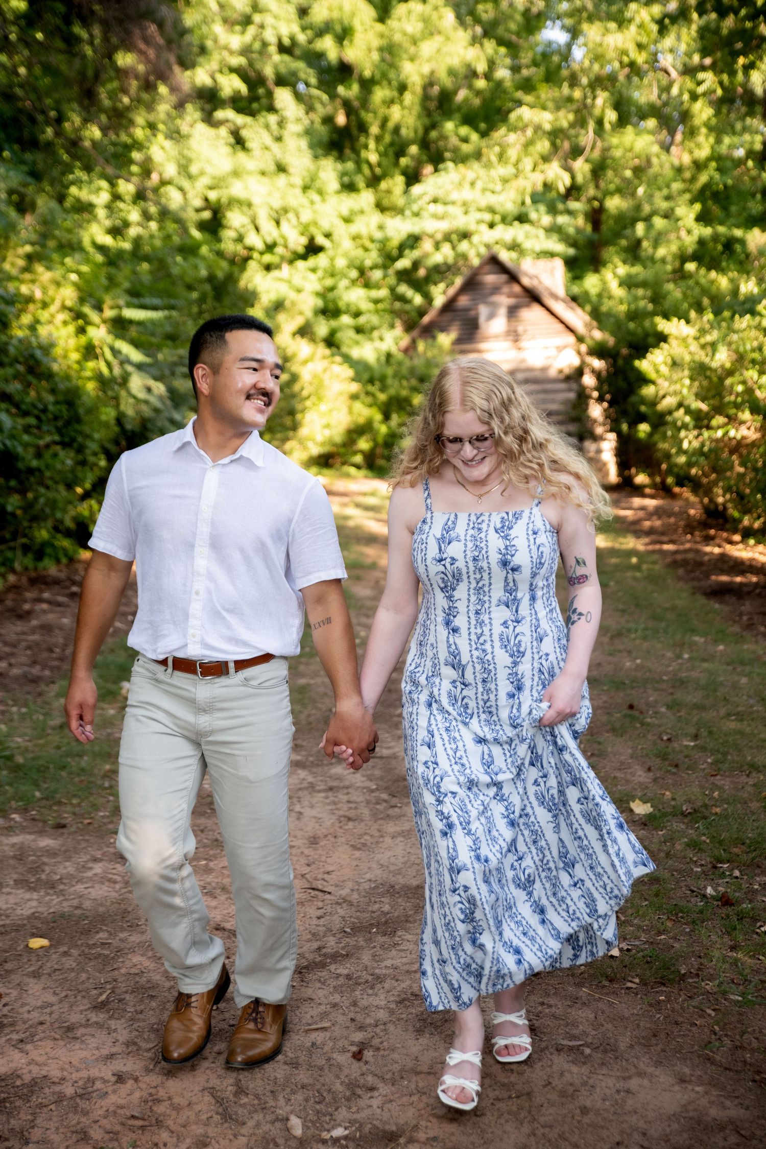 A couple walks hand-in-hand on a dirt path surrounded by lush greenery, smiling at each other.