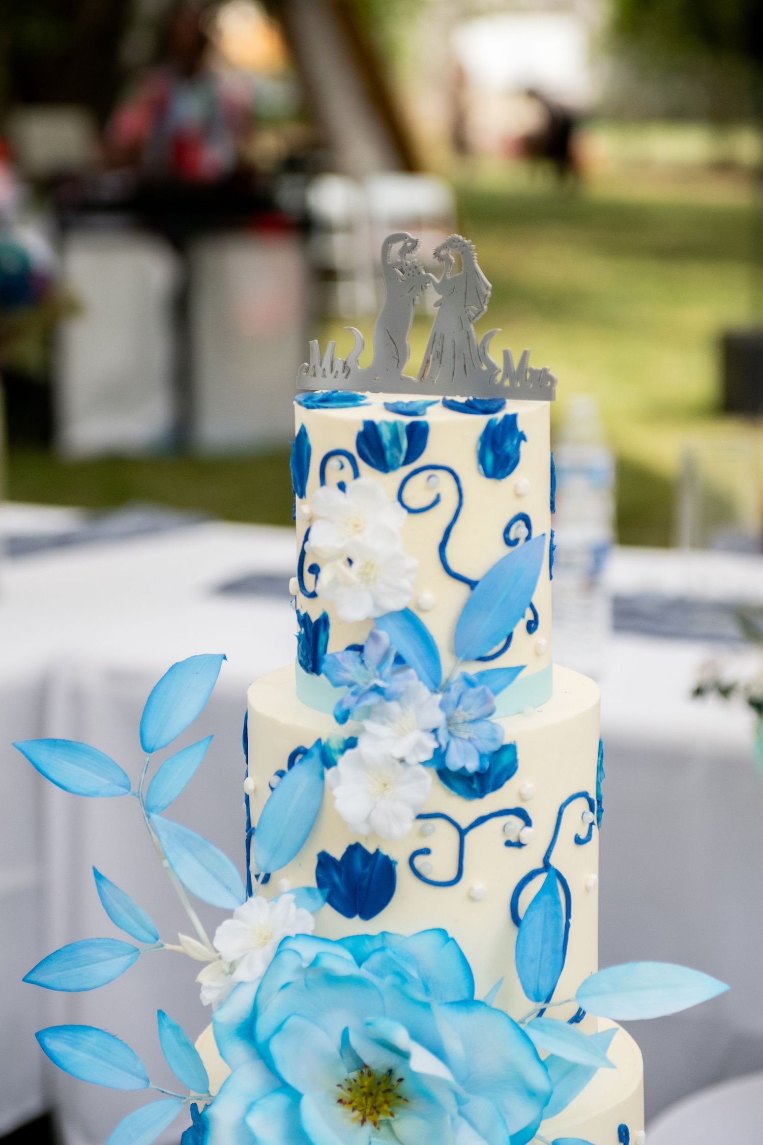 Elegant three-tier white wedding cake with blue floral designs and Mr. & Mrs. topper at outdoor reception.