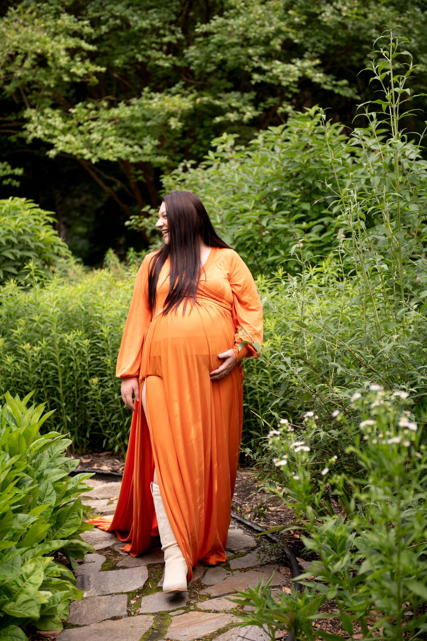 Pregnant woman in orange maxi dress walking on stone path in lush green garden, cradling baby bump.