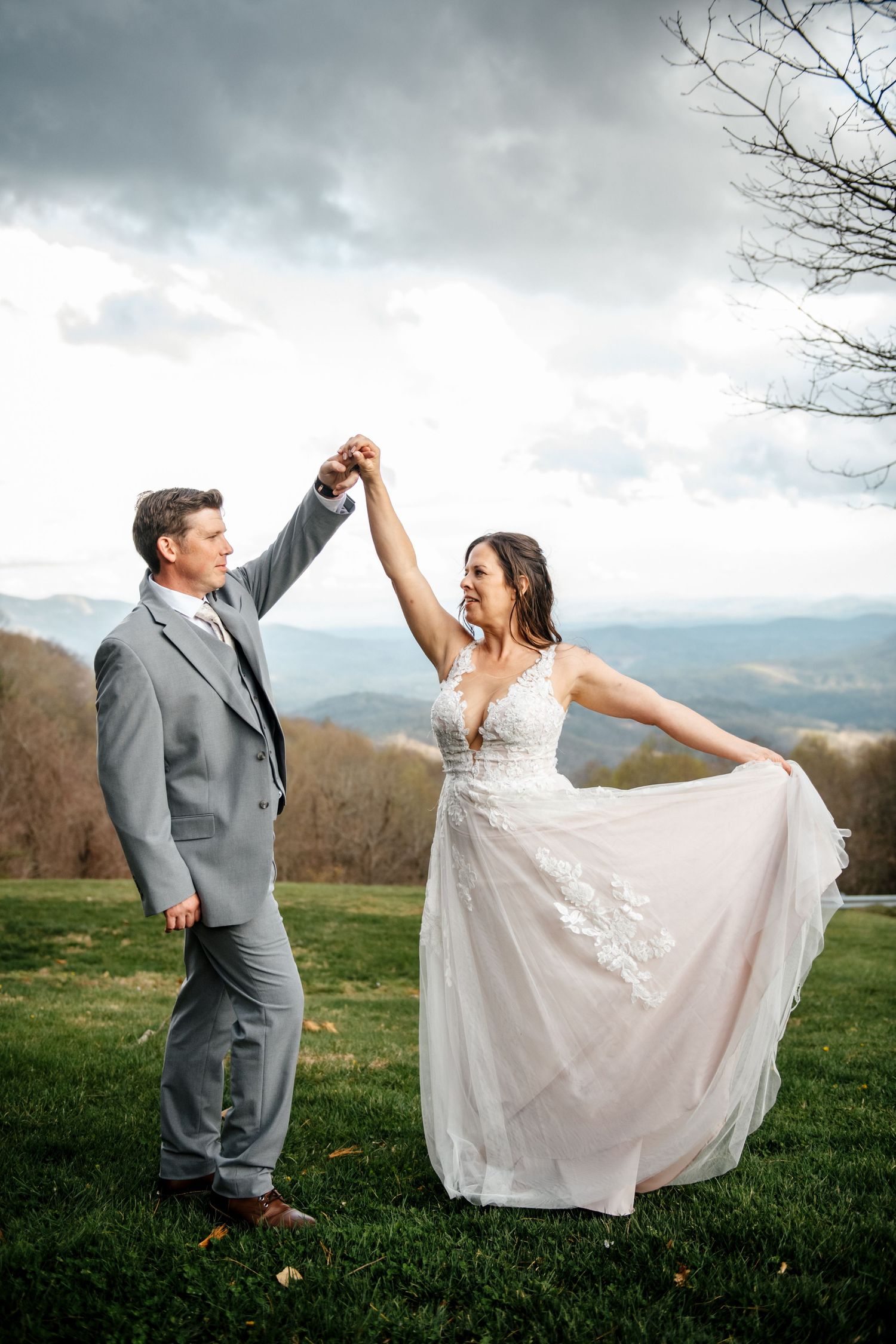 Bride and groom dancing outdoors on green grass with mountain views and cloudy sky in wedding attire.