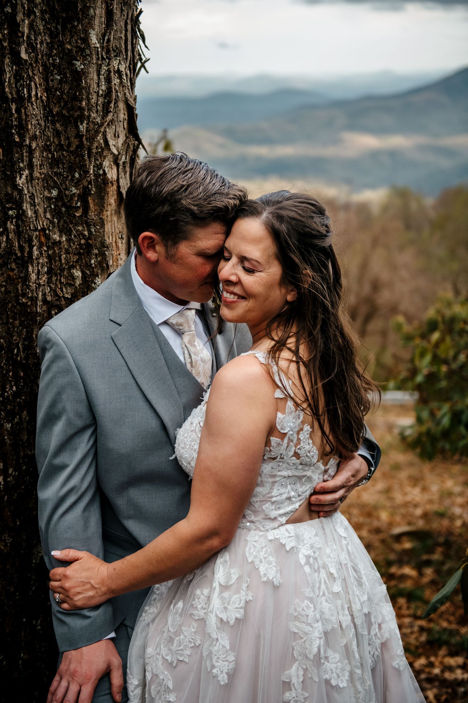 A couple in wedding attire embraces near a tree with scenic mountain views in the background.