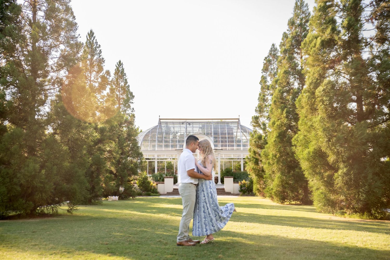 Couple embracing on green lawn before glass greenhouse surrounded by tall trees in golden sunlight.