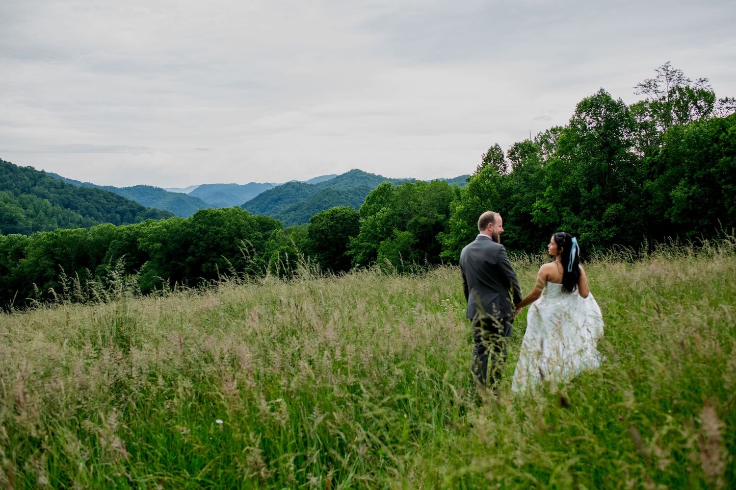 Bride and groom holding hands in a lush green meadow with rolling mountain views on their wedding day.