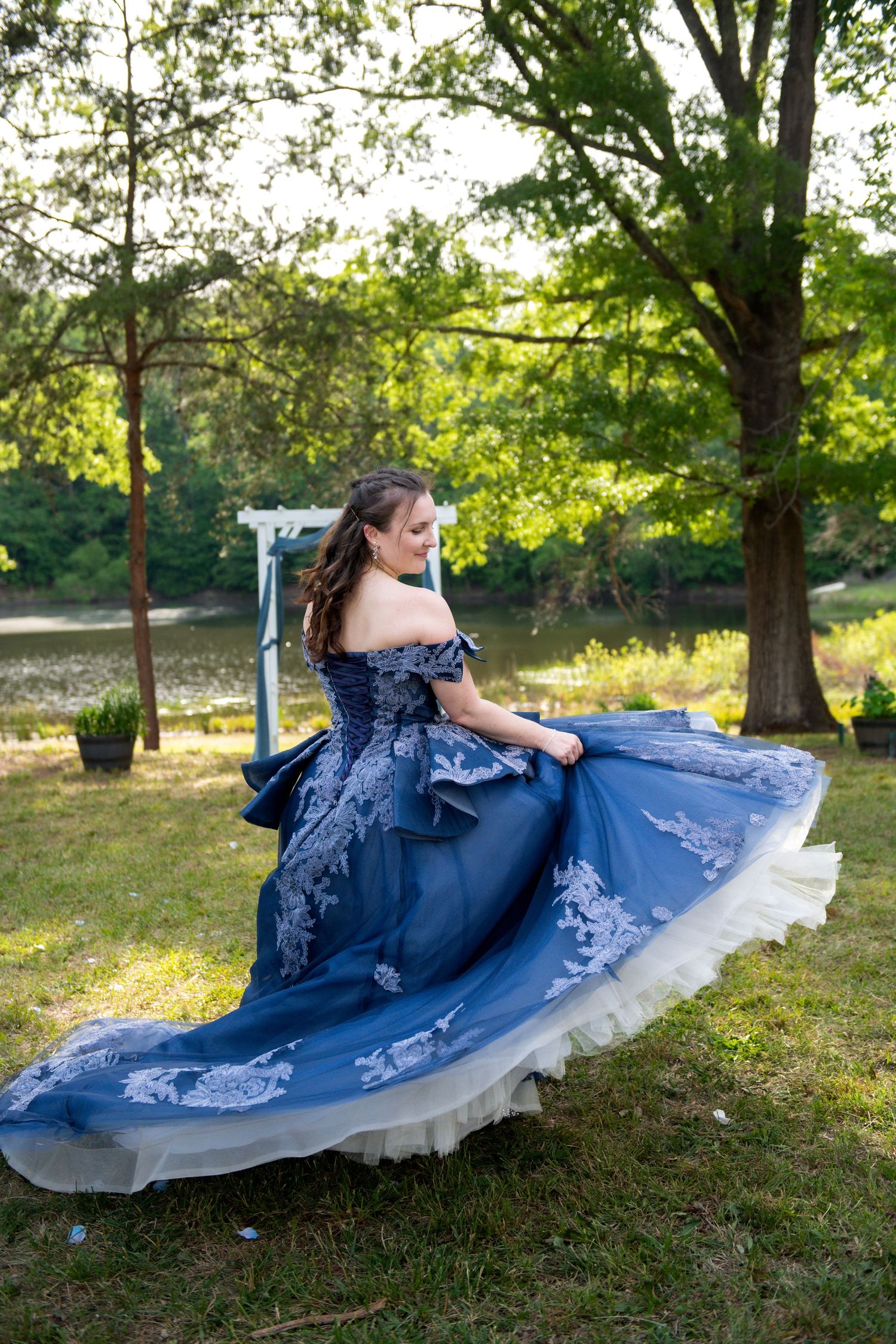 Young woman in elegant blue off-shoulder ball gown with white lace twirling outdoors near a lake.