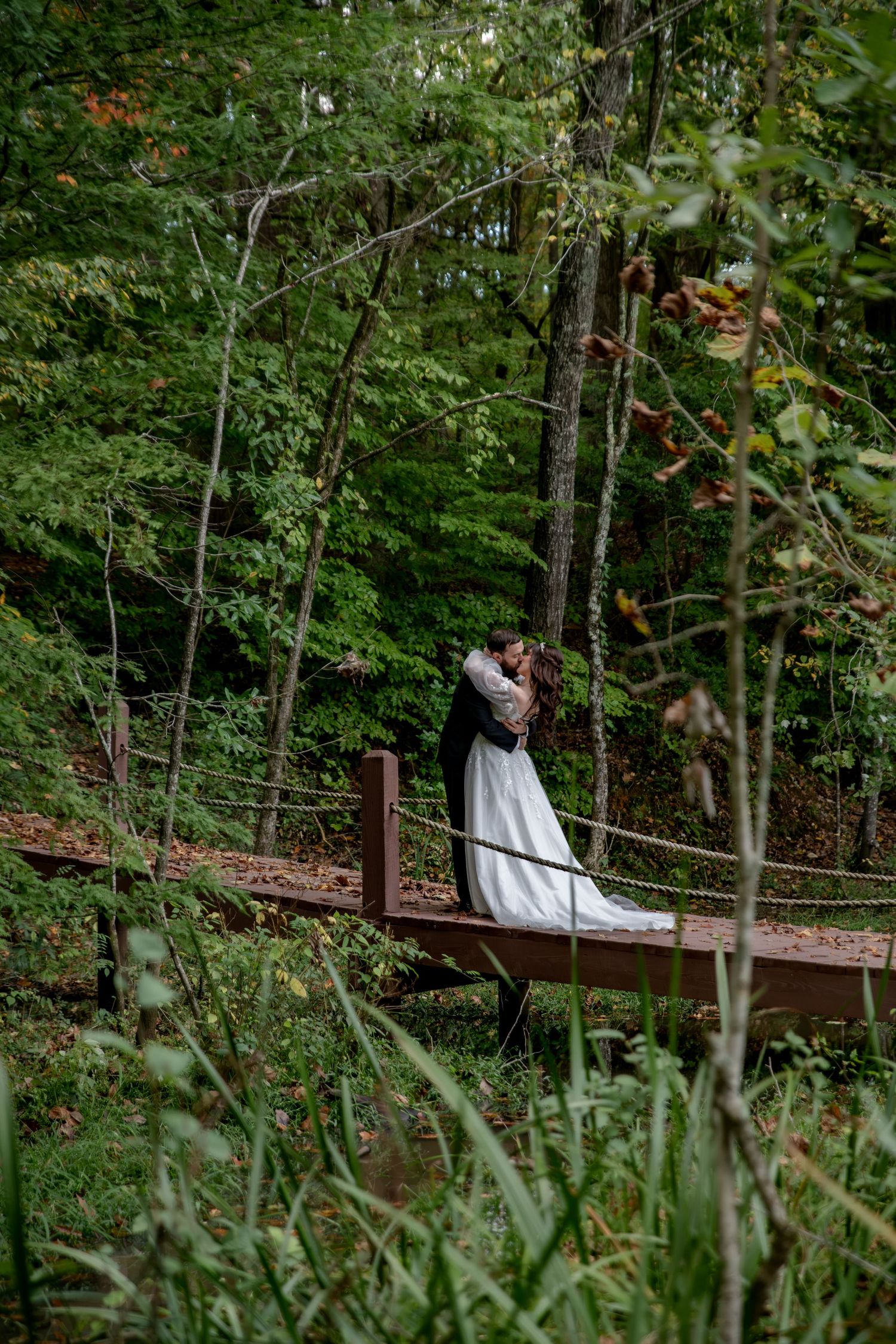 Bride in a white gown and groom in a dark suit embrace on a wooden footbridge surrounded by lush green forest trees.