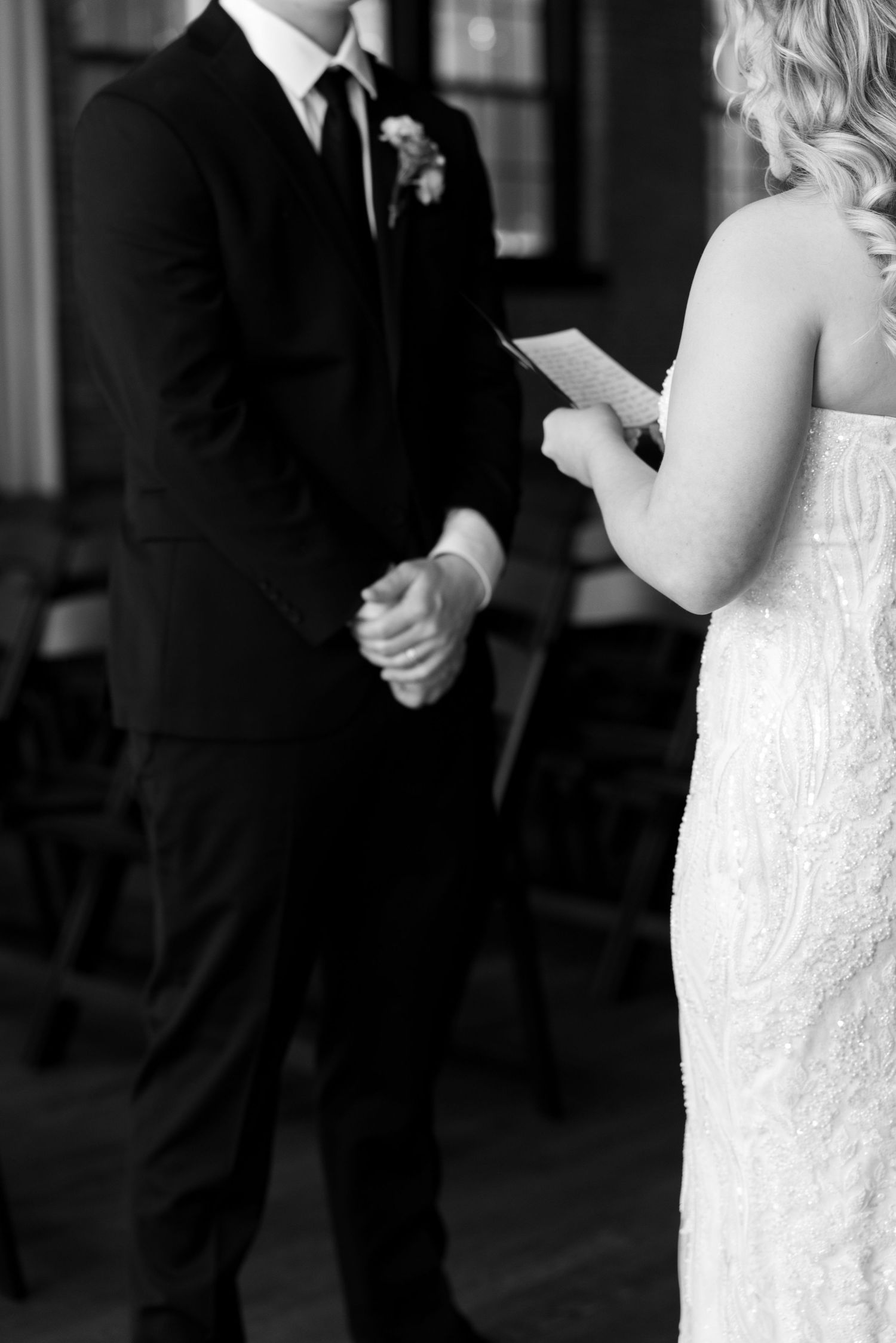 Black and white photo of bride reading wedding vows while holding hands with groom during indoor ceremony.