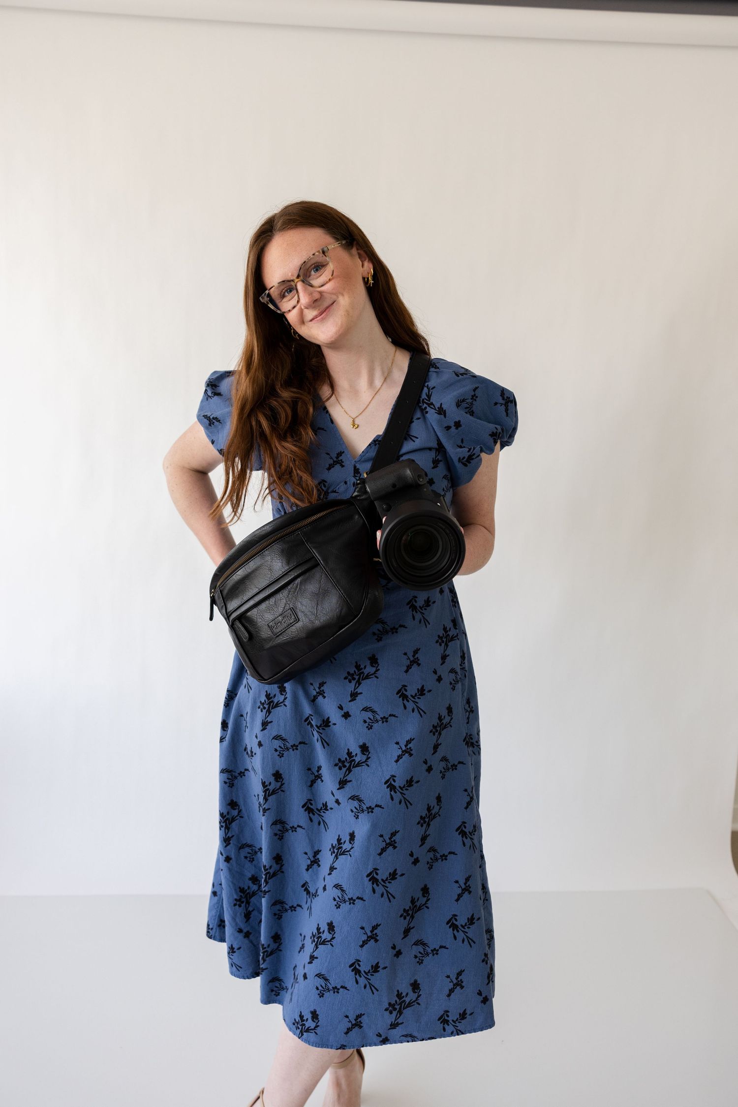 Woman in blue floral midi dress with puff sleeves wearing glasses and black leather crossbody bag, smiling.