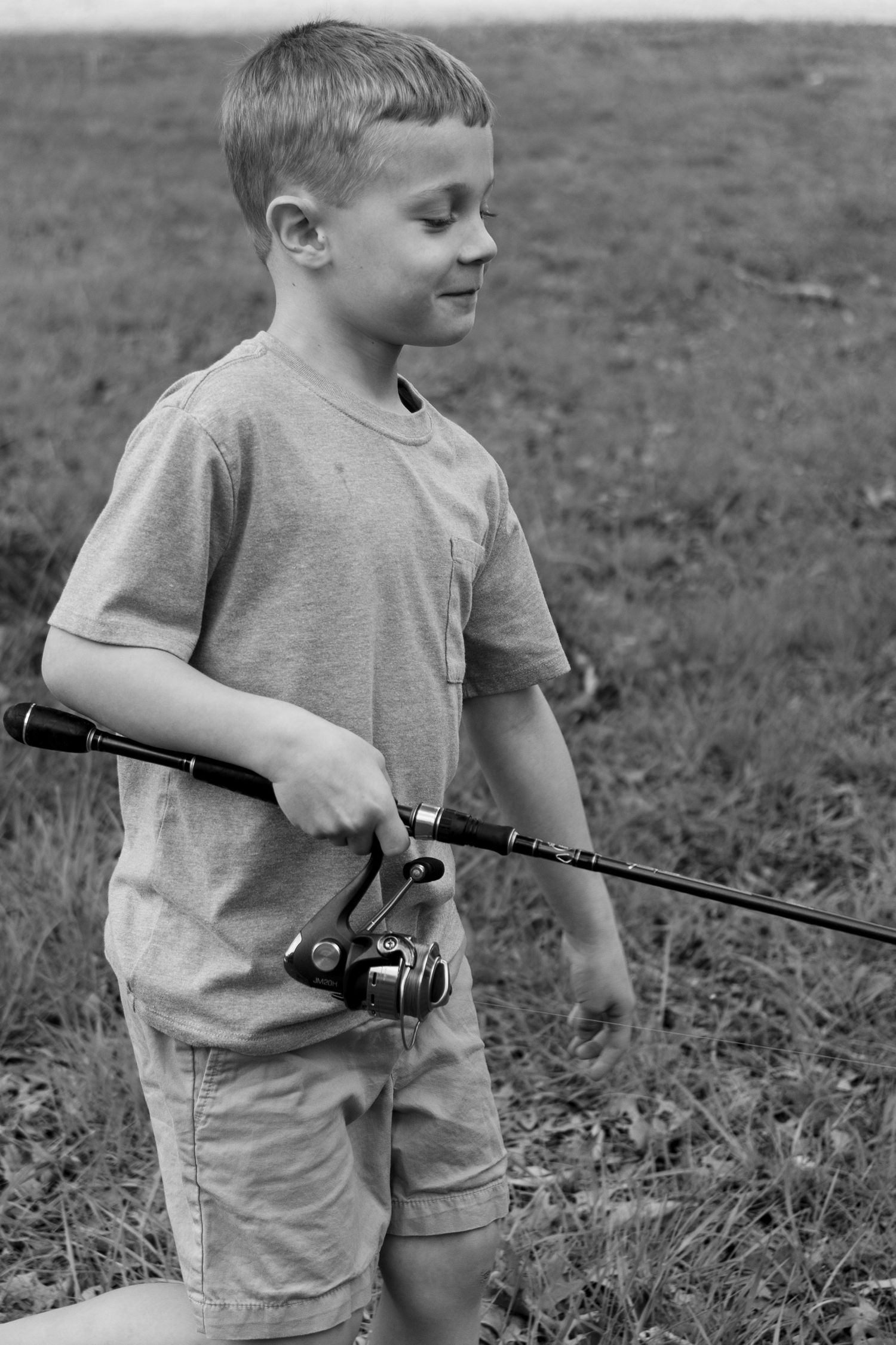 Young child in casual attire holds a fishing rod while standing in a grassy field in black and white.