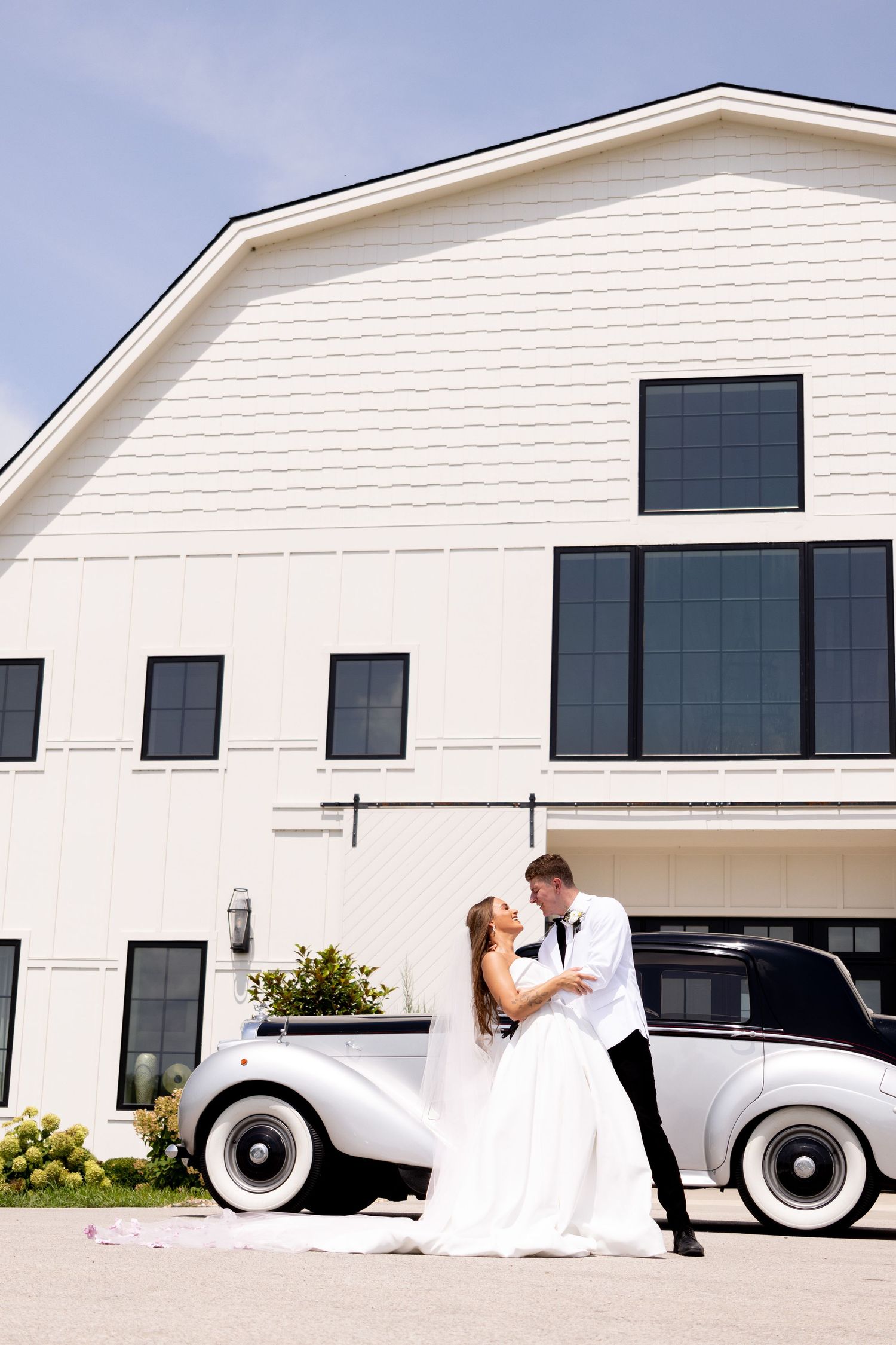 Bride and groom share a romantic kiss beside a vintage white classic car in front of an elegant white barn venue.
