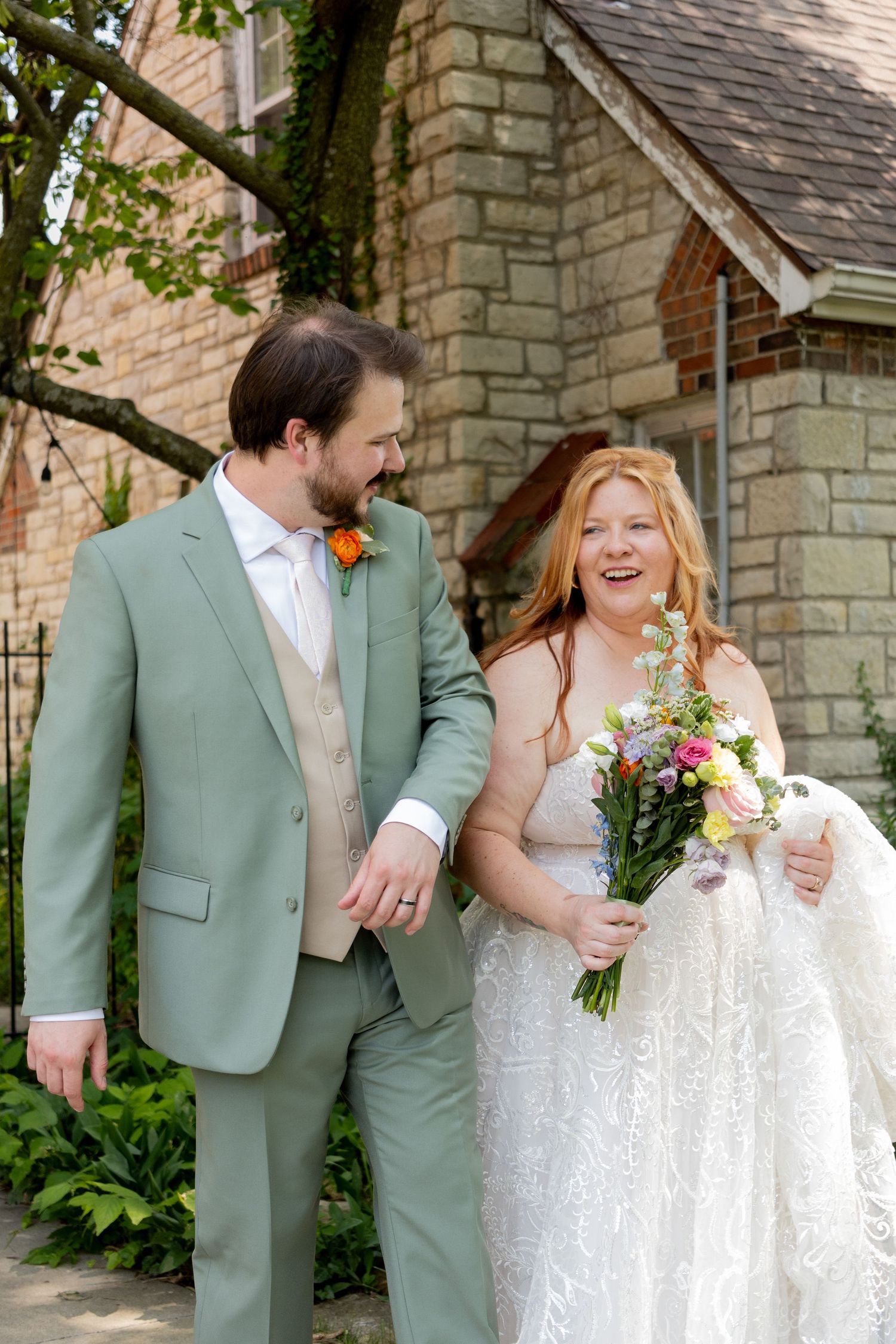 Couple in wedding attire share joyful moment outside stone building.