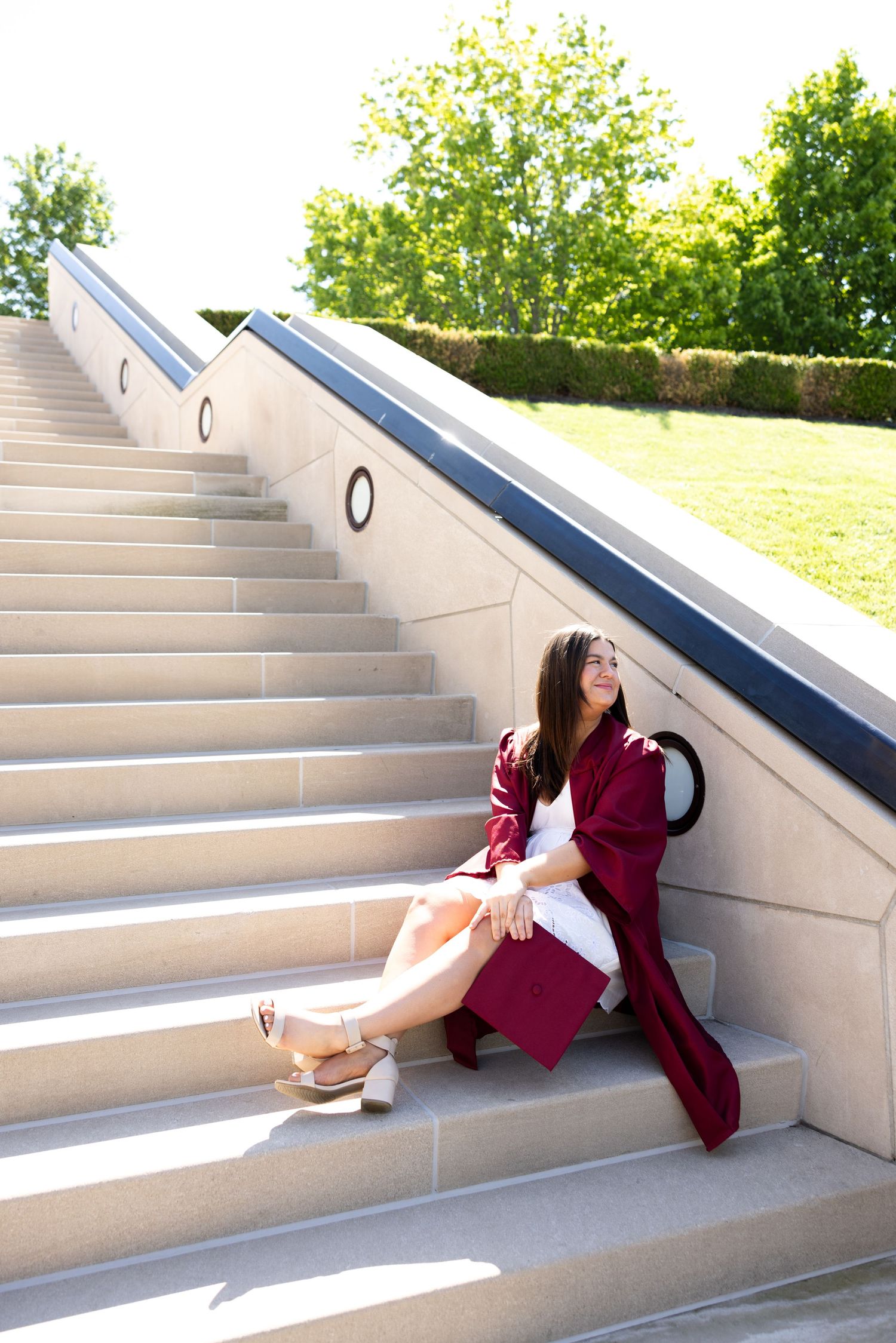 Young woman in maroon graduation gown sitting on outdoor stone steps, smiling, with green trees in background.