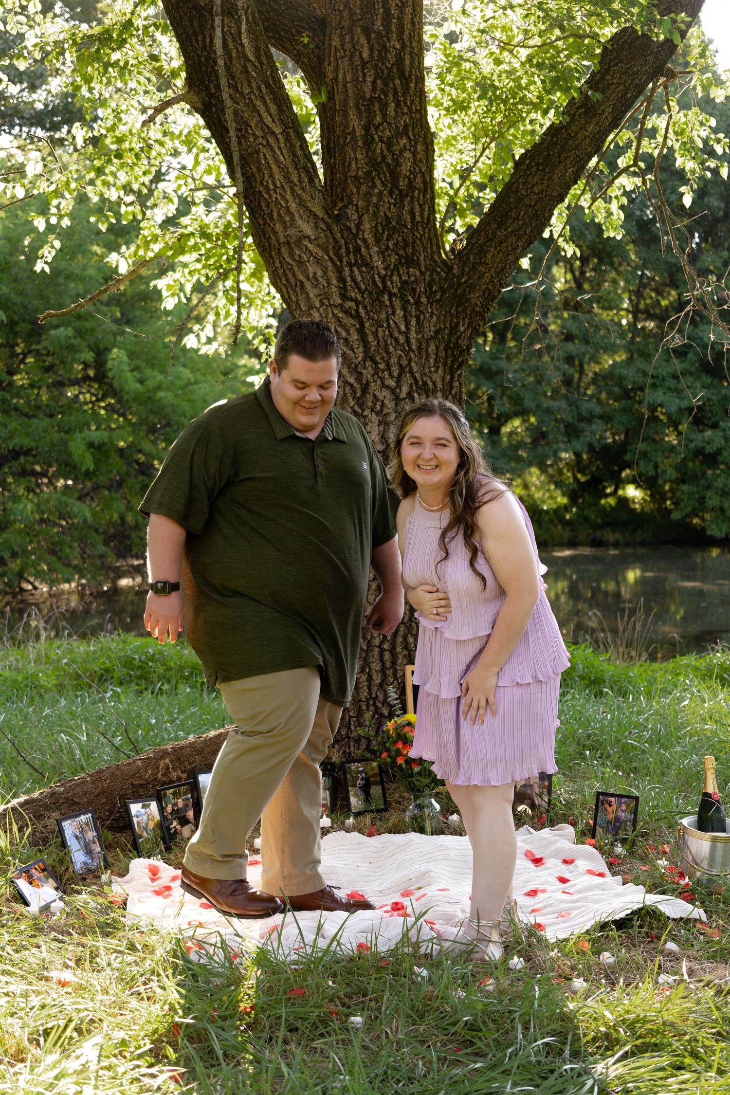 A couple sits together on a picnic blanket under a large tree in a sunny garden setting.
