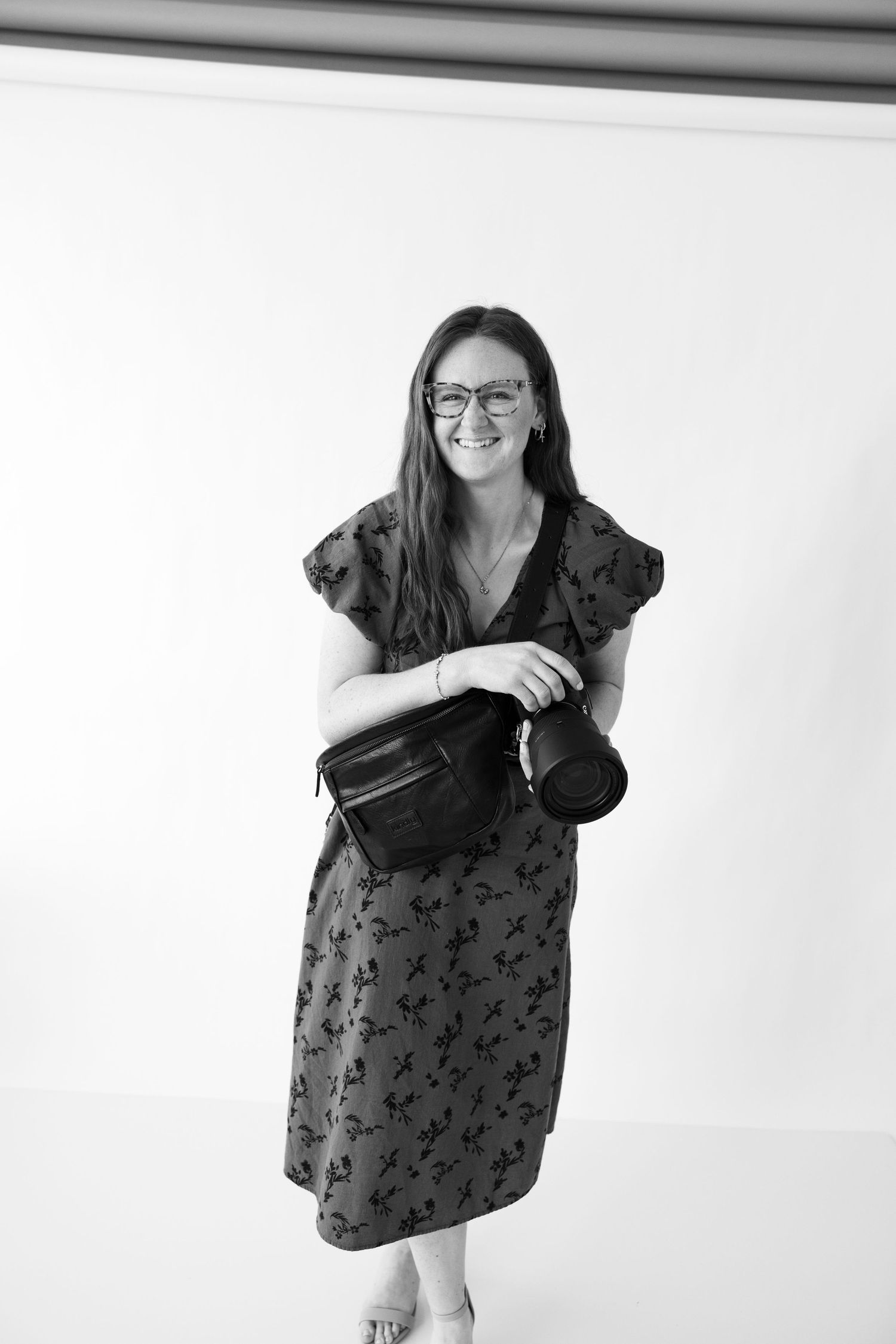 Smiling woman with glasses in floral dress holding a camera, black and white portrait photo.