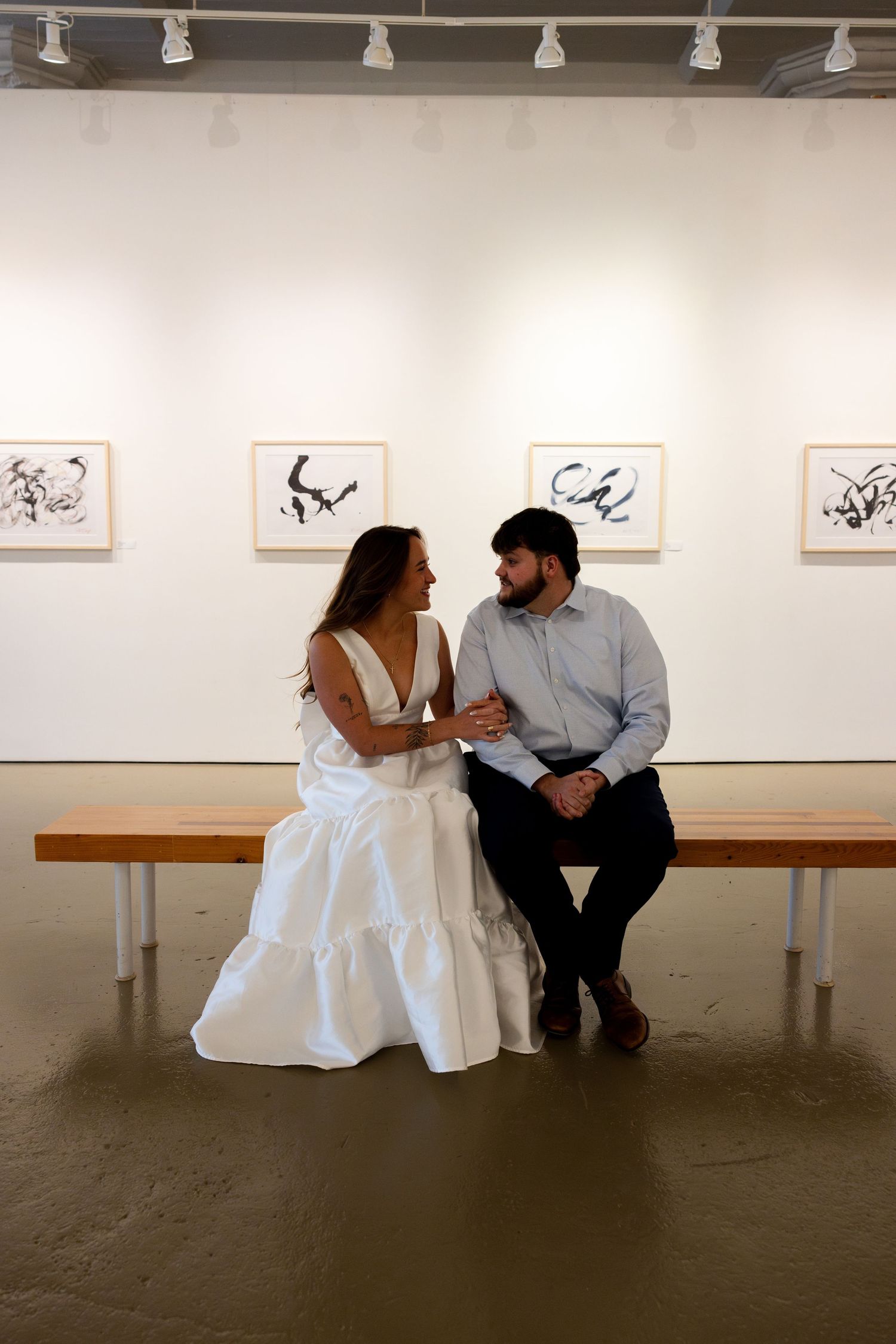 A couple sits together on a bench in an art gallery with framed artwork displayed on white walls behind them.