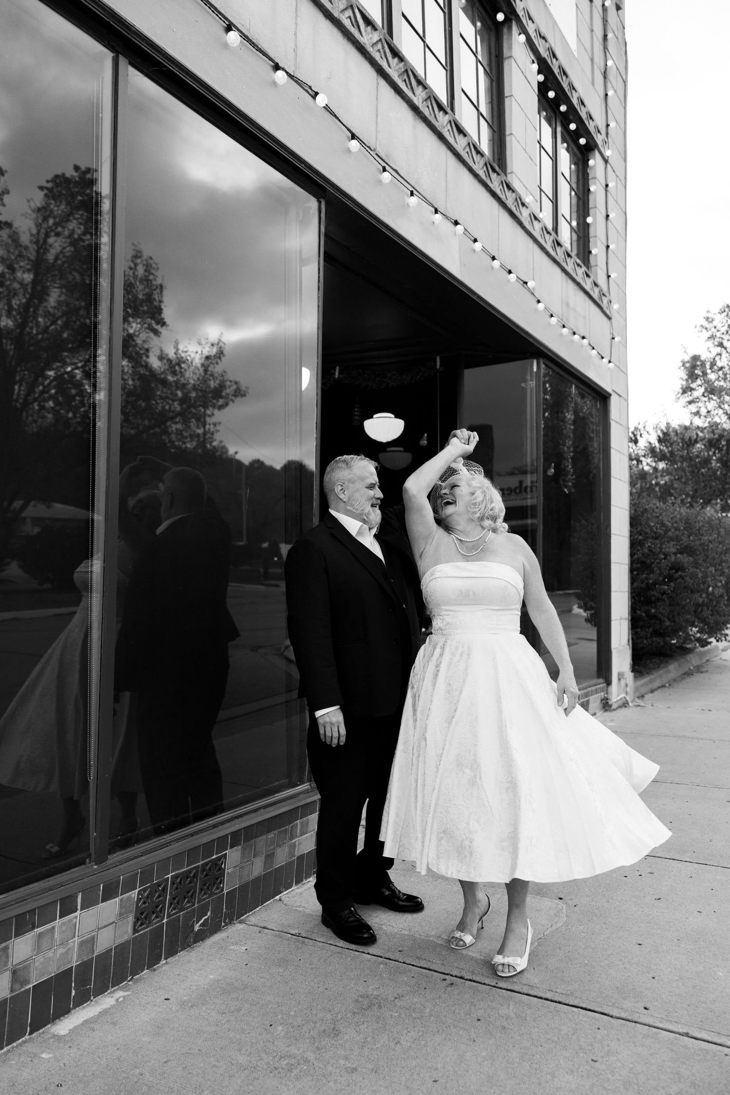 Black and white photo of a bride and groom dancing outside a building, bride twirling in a tea-length wedding dress.