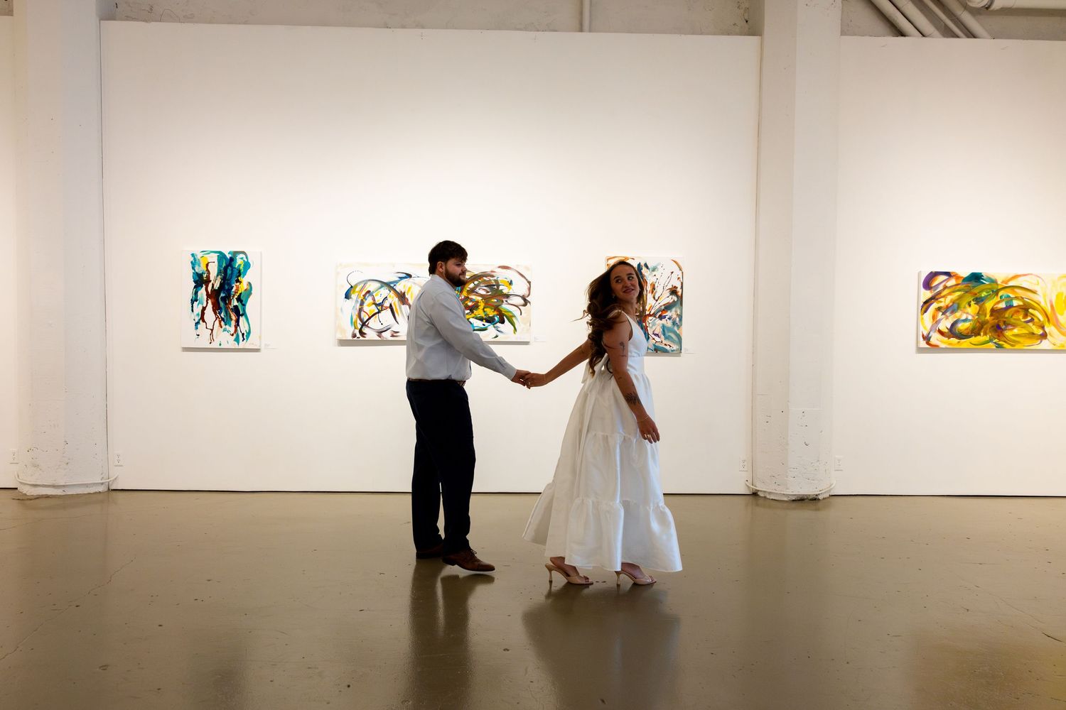 A couple dances in an elegant white art gallery with paintings displayed on the walls.