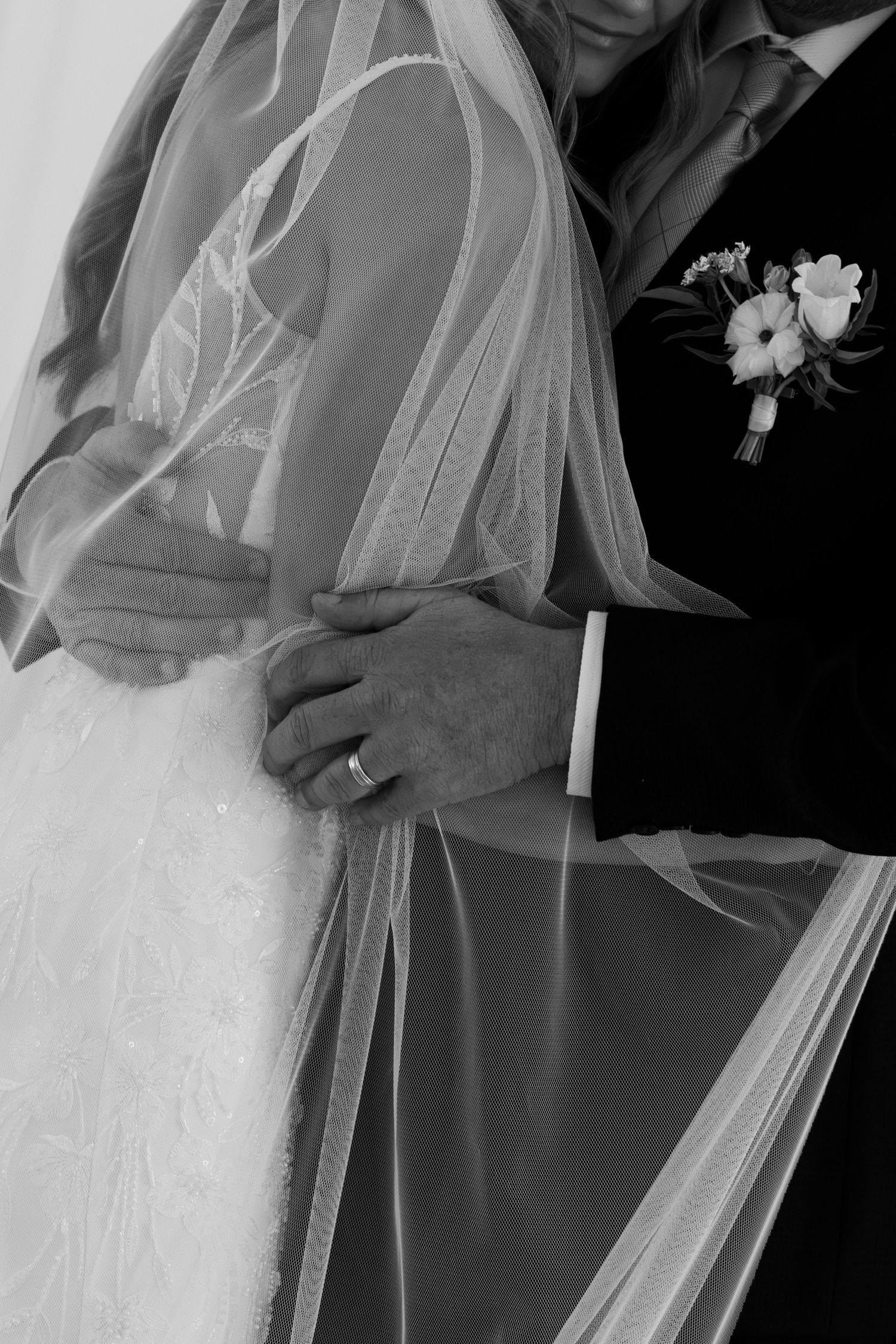 Black and white wedding photo of bride and groom embracing, showing rings, veil, and boutonniere.